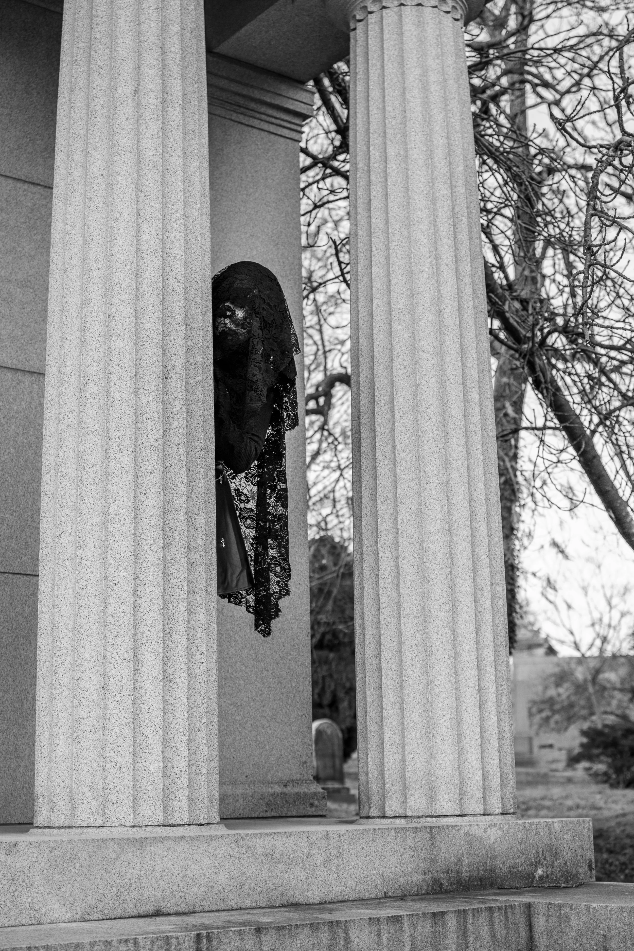 A woman wearing a lace head covering and dress is kneeling behind the columns of a stone structure, possibly a mausoleum or monument, with leafless trees in the background.