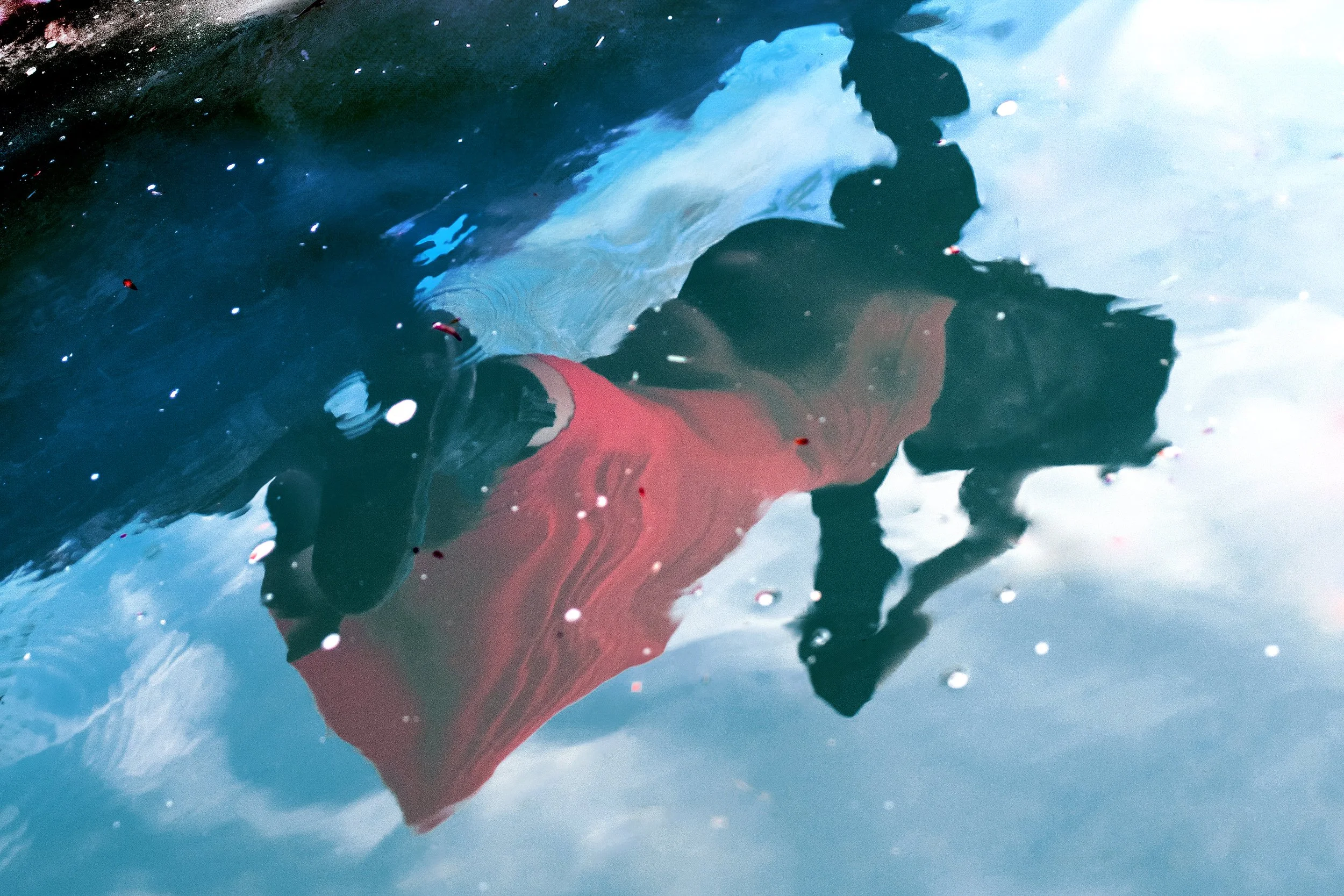A person in a red shirt and black pants is swimming underwater in a pool, with their face and upper body visible through the water's surface.