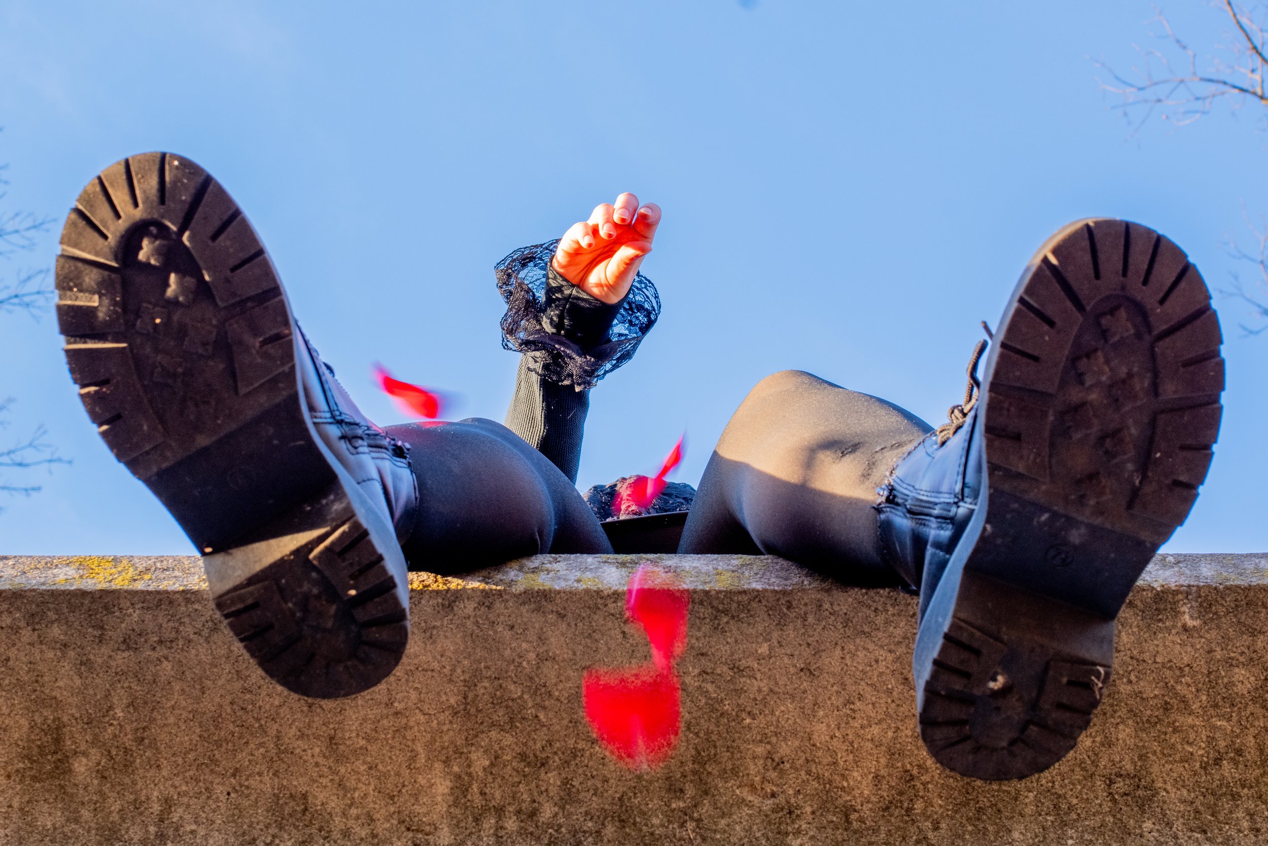 Person sitting on a brick wall with their legs stretched out, wearing black shoes and beige pants, holding their hand up with fingers in a gesture, against a clear blue sky.