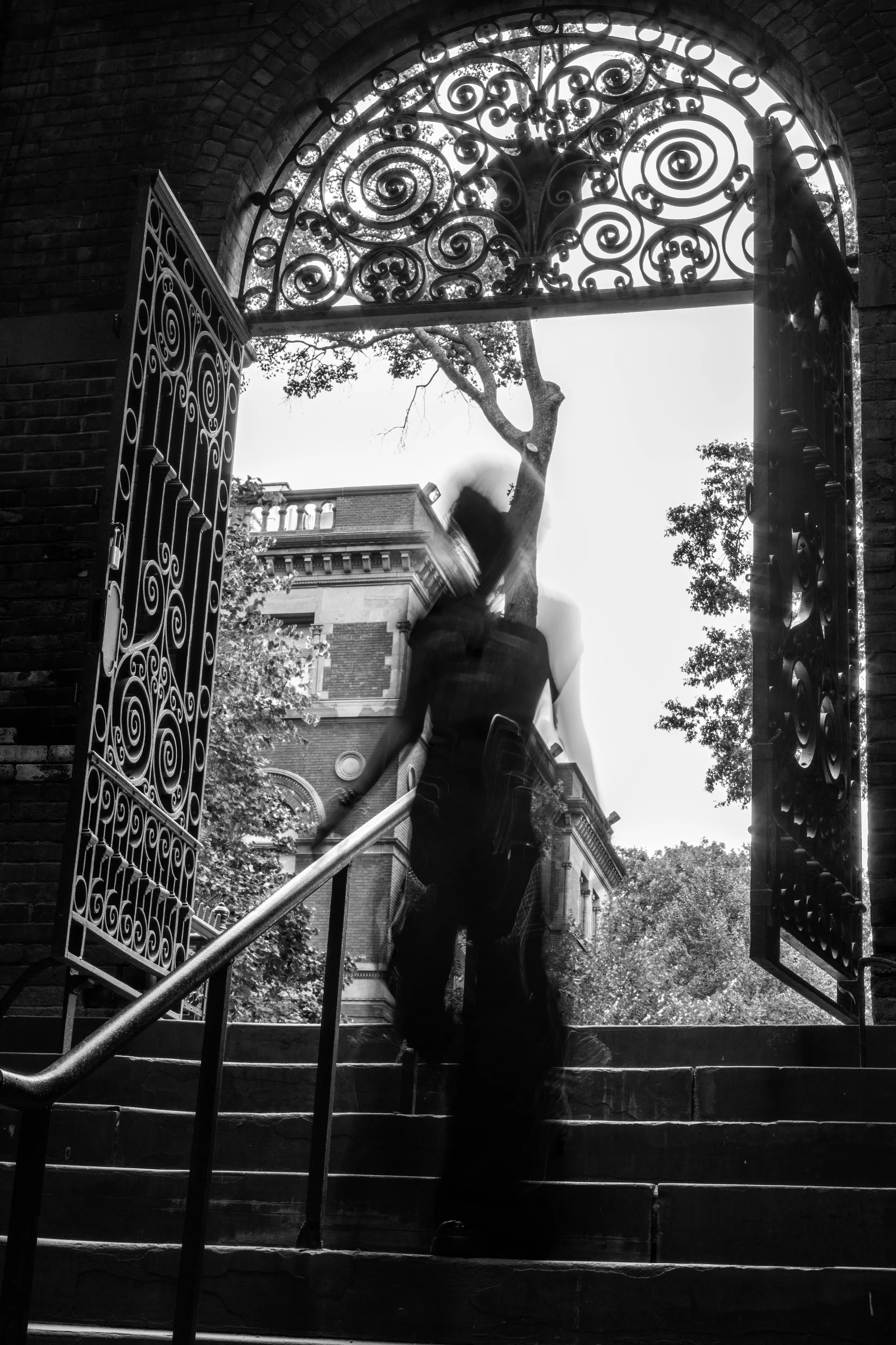 A person ascending stairs through an ornate, wrought-iron gate with intricate swirling patterns, framed by brick arches and trees in the background, in a black and white photo.