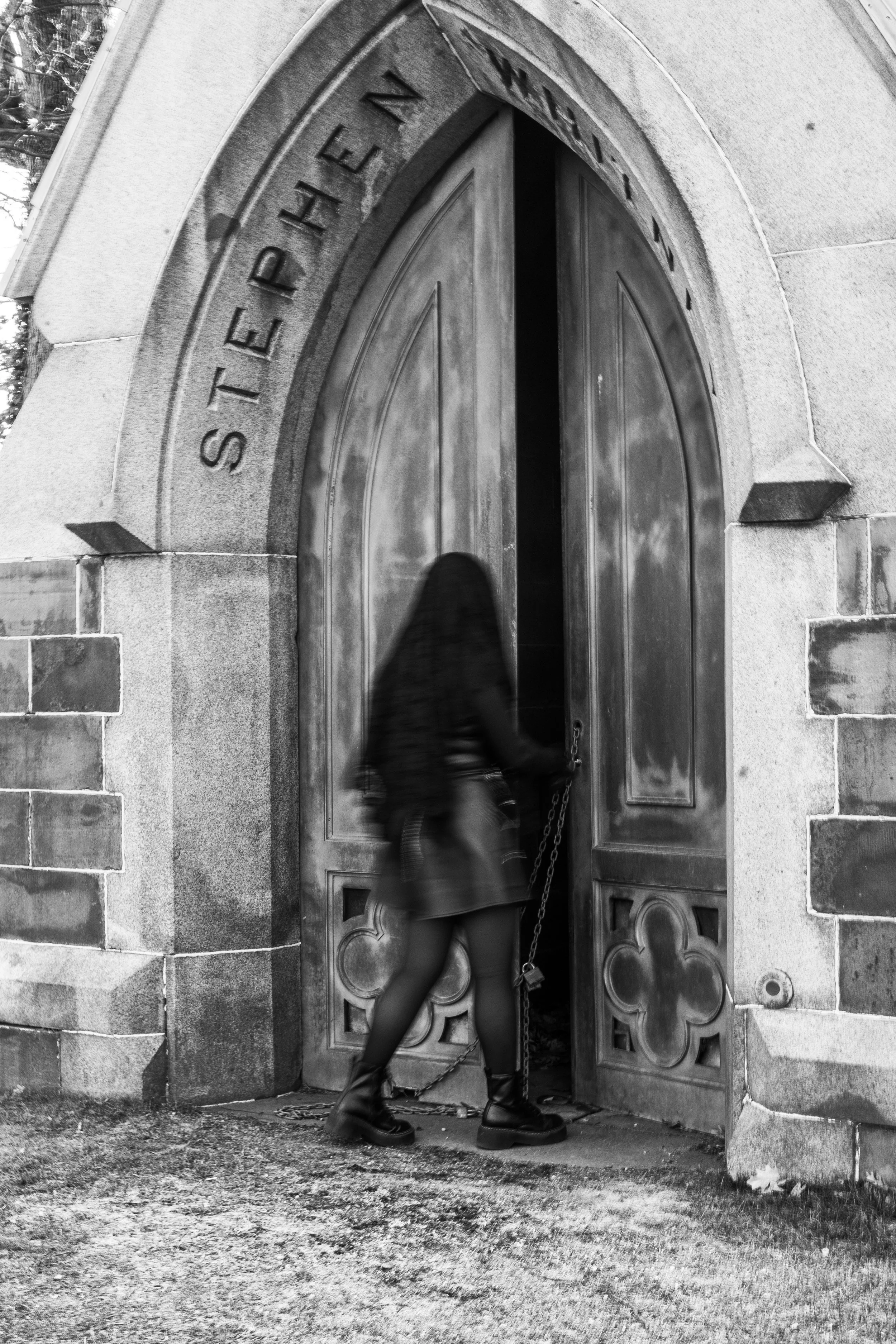Black and white photo of a person with long hair and a skirt walking through an arched gate with inscribed text, holding a chain.