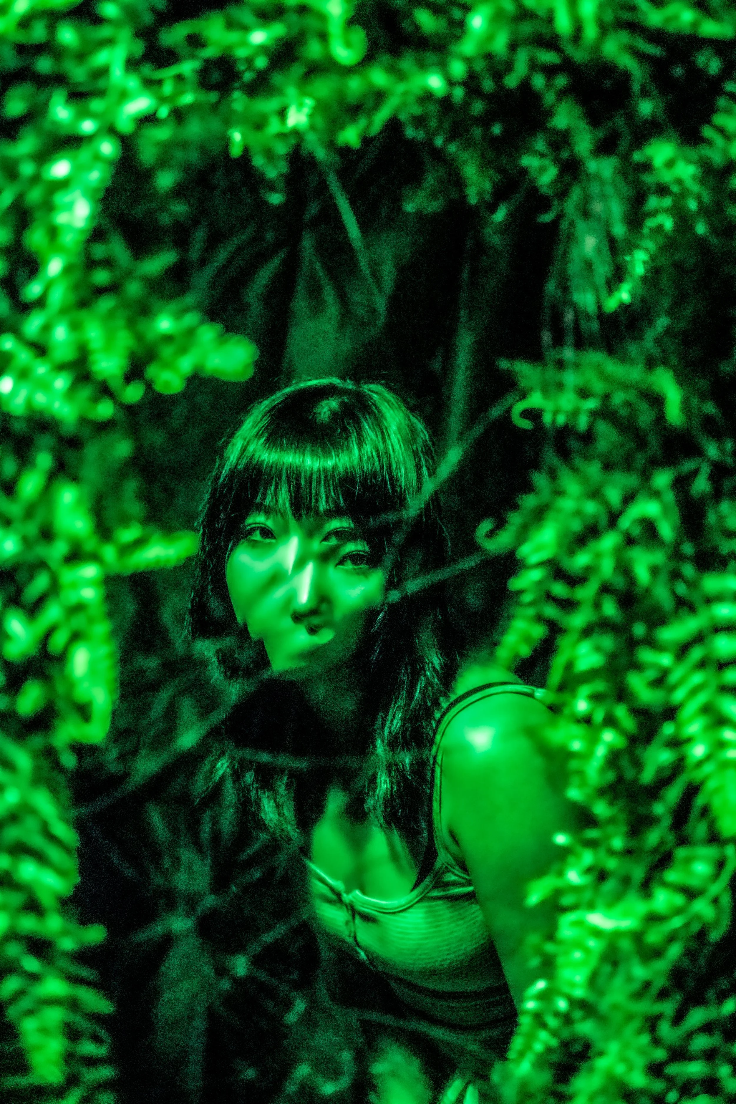 The image shows a woman with long hair and bangs taking a selfie through a leafy plant in a green-lit environment.