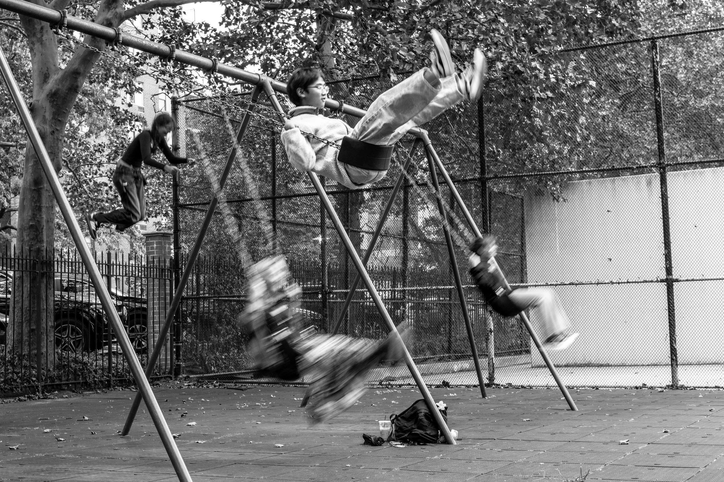 Children playing on swings at a playground enclosed by a chain-link fence, with trees and parked cars in the background.