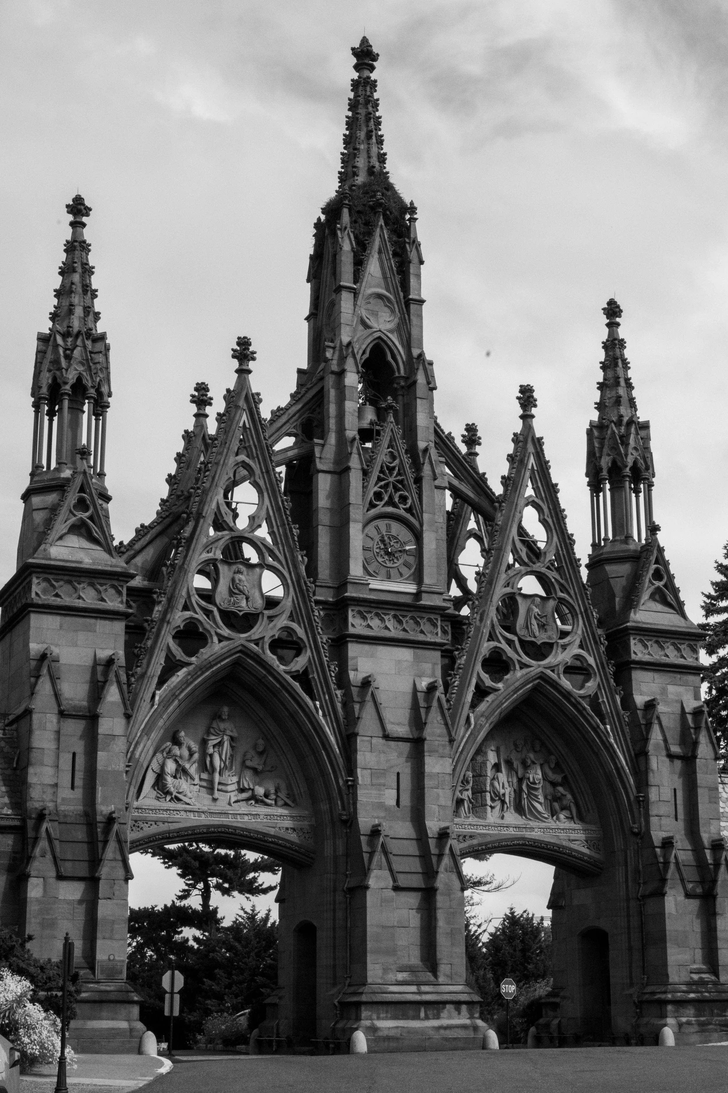 Black and white photo of a Gothic-style cathedral facade with ornate spires and sculptures, topped by a clock, framed by trees and street elements.