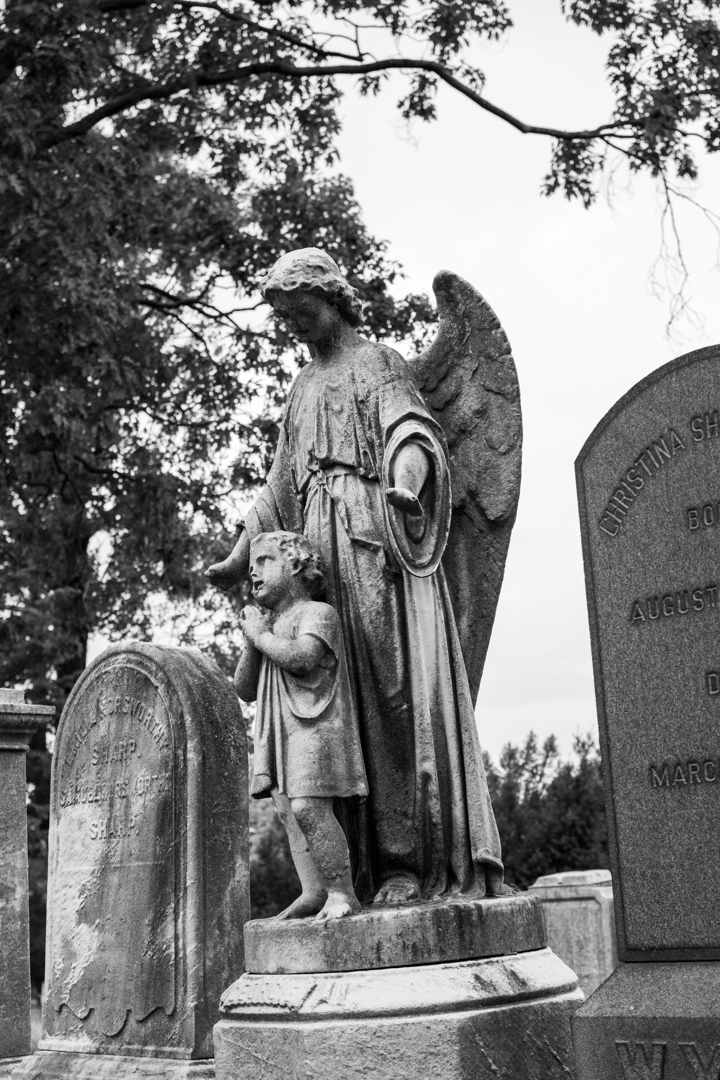 Black and white photograph of a stone angel statue in a cemetery, standing next to a gravestone, with trees in the background.