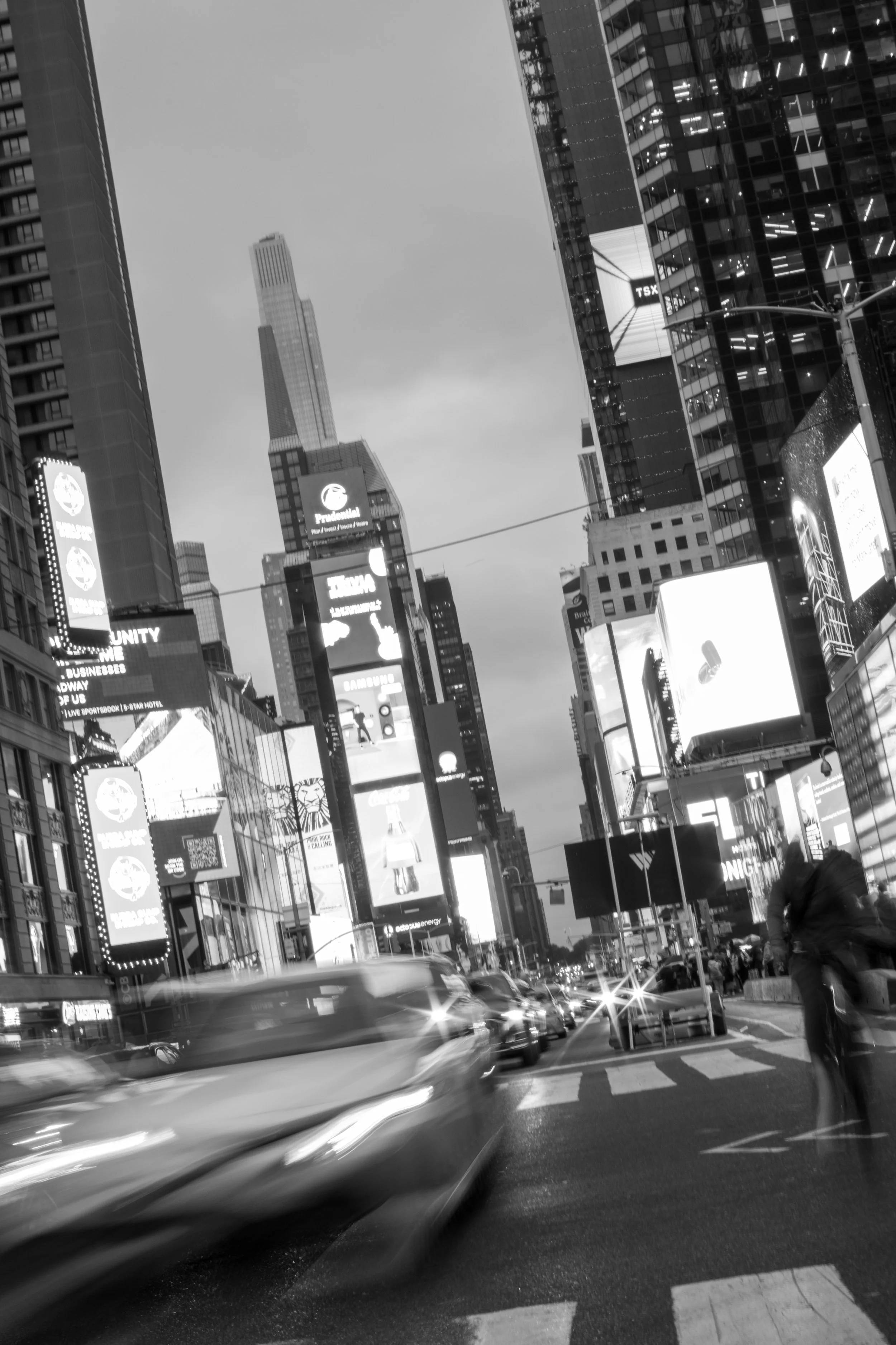 Black and white photo of Times Square in New York City with tall buildings, billboards, and a busy street with moving cars and pedestrians.