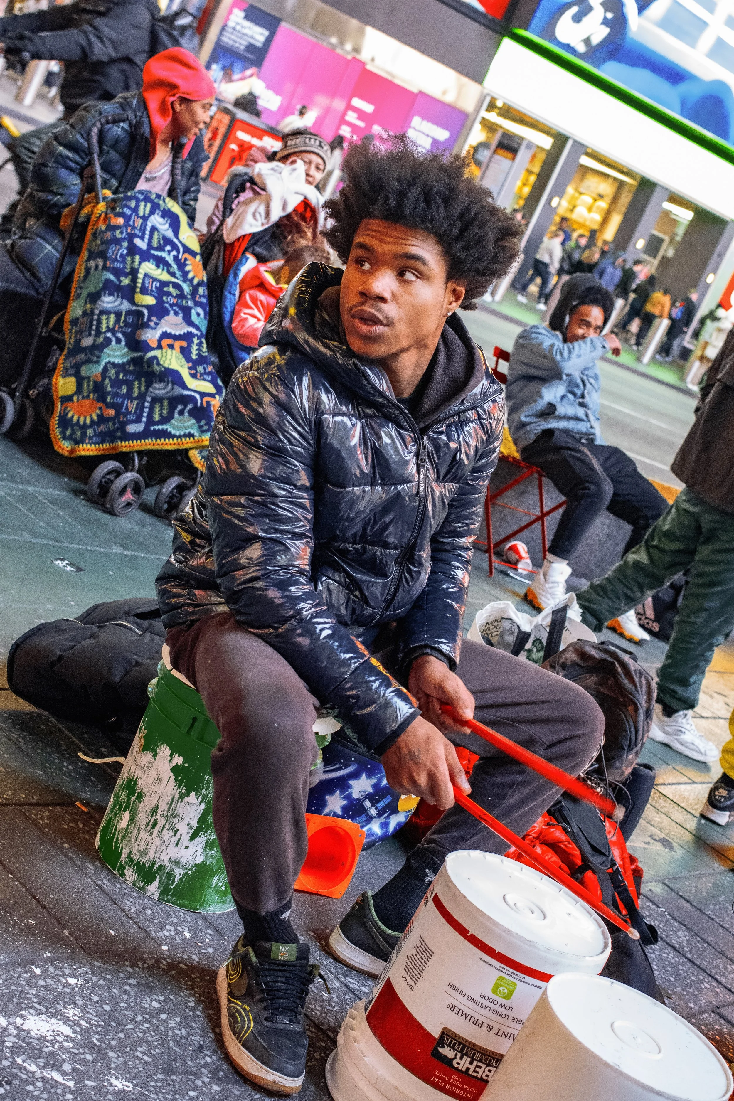 Young man with an afro hairstyle wearing a shiny black puffer jacket sitting on a small cooler on a city sidewalk, holding a red stick, with buckets, bags, and a colorful skateboard around him. In the background, several other people are sitting and 