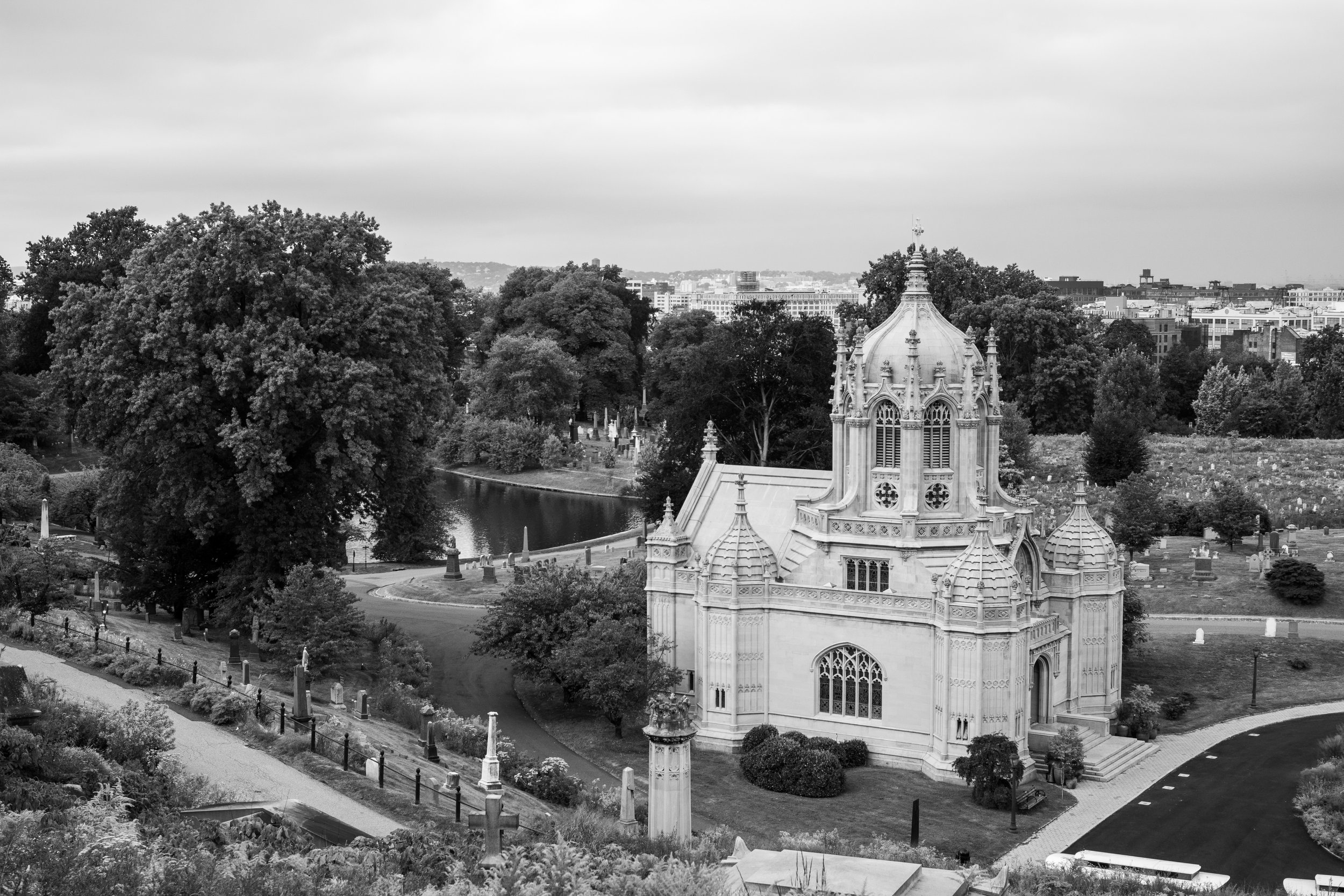 A black-and-white photo of a historic church with ornate architecture, surrounded by a graveyard with tombstones, trees, and a paved path, with a city skyline in the background.