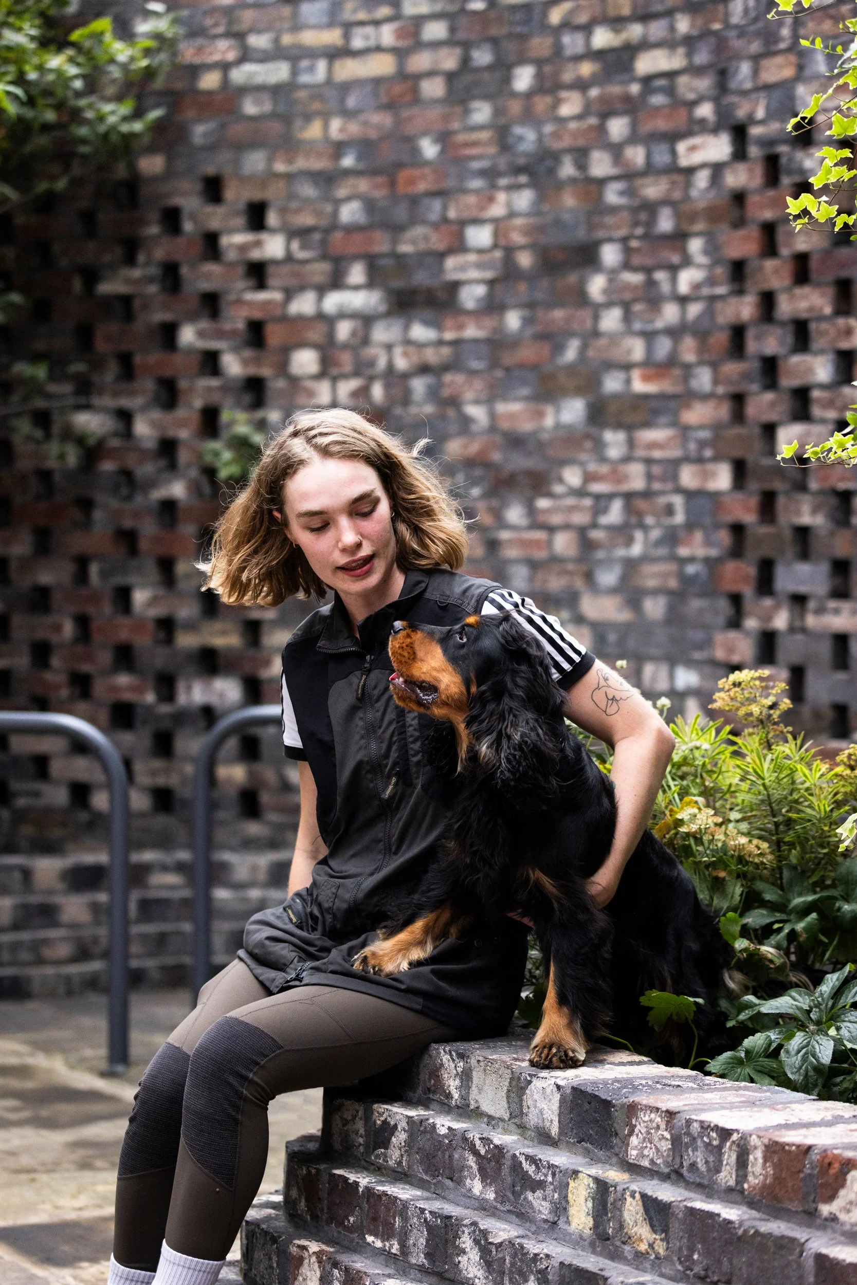 A young woman with shoulder-length blonde hair, wearing a black vest and brown riding pants, sits on a brick wall. She is petting and looking at ablack and tan dog, sitting on the wall beside her. The background features a red brick wall with some gr