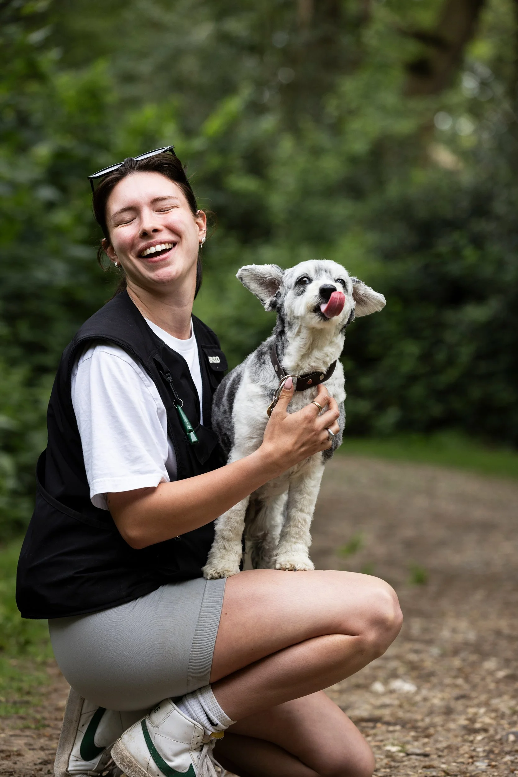 A smiling woman kneeling on a dirt path in a wooded area, holding a happy dog with its tongue out.