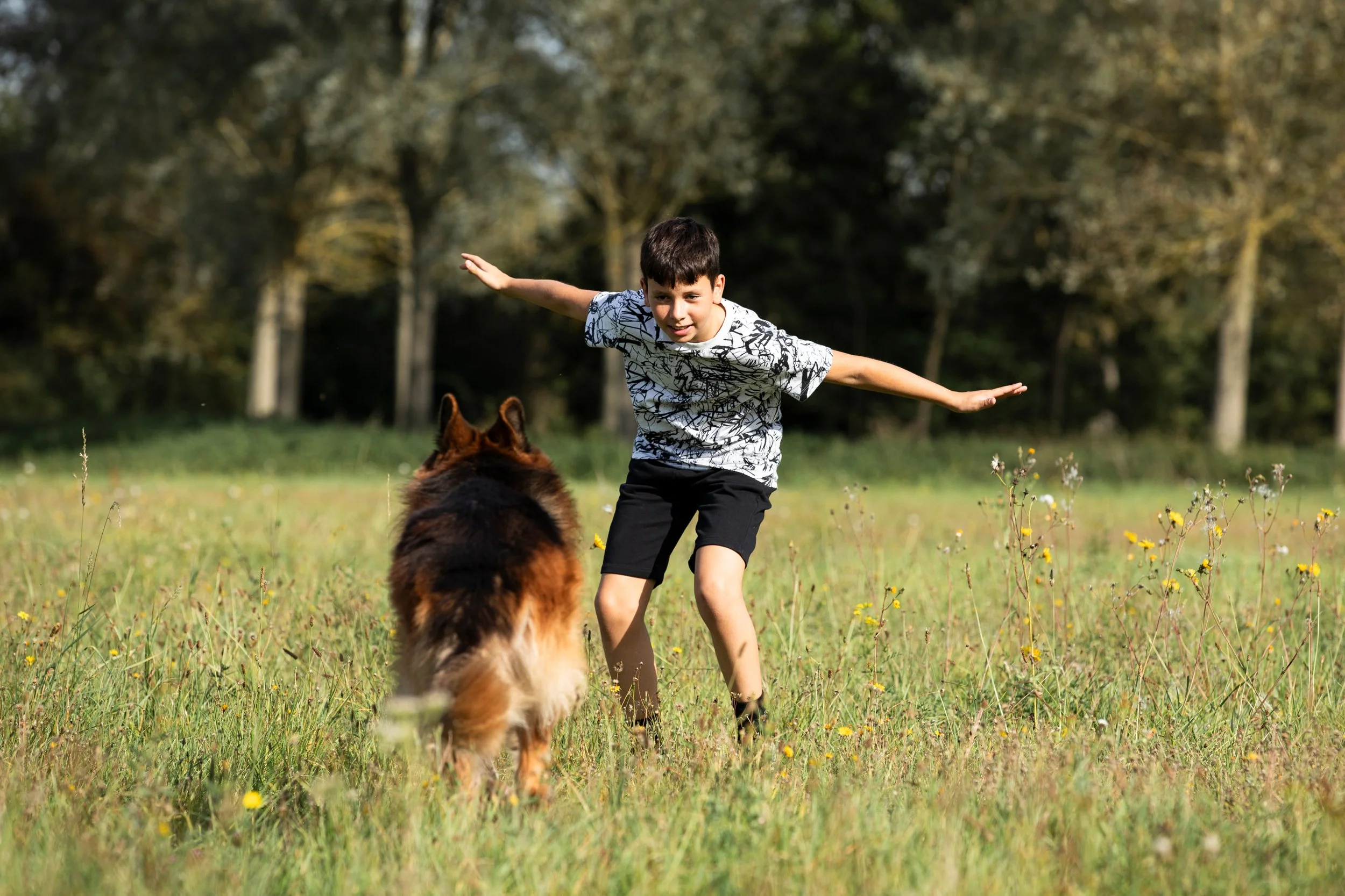 A boy with outstretched arms playing with a large dog in a grassy field during daytime.