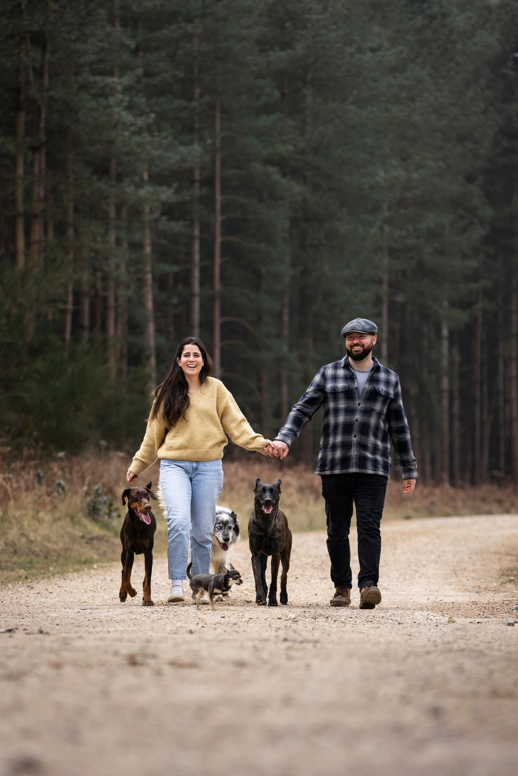 A couple walking with four dogs on a dirt path in a forested area, holding hands and smiling.