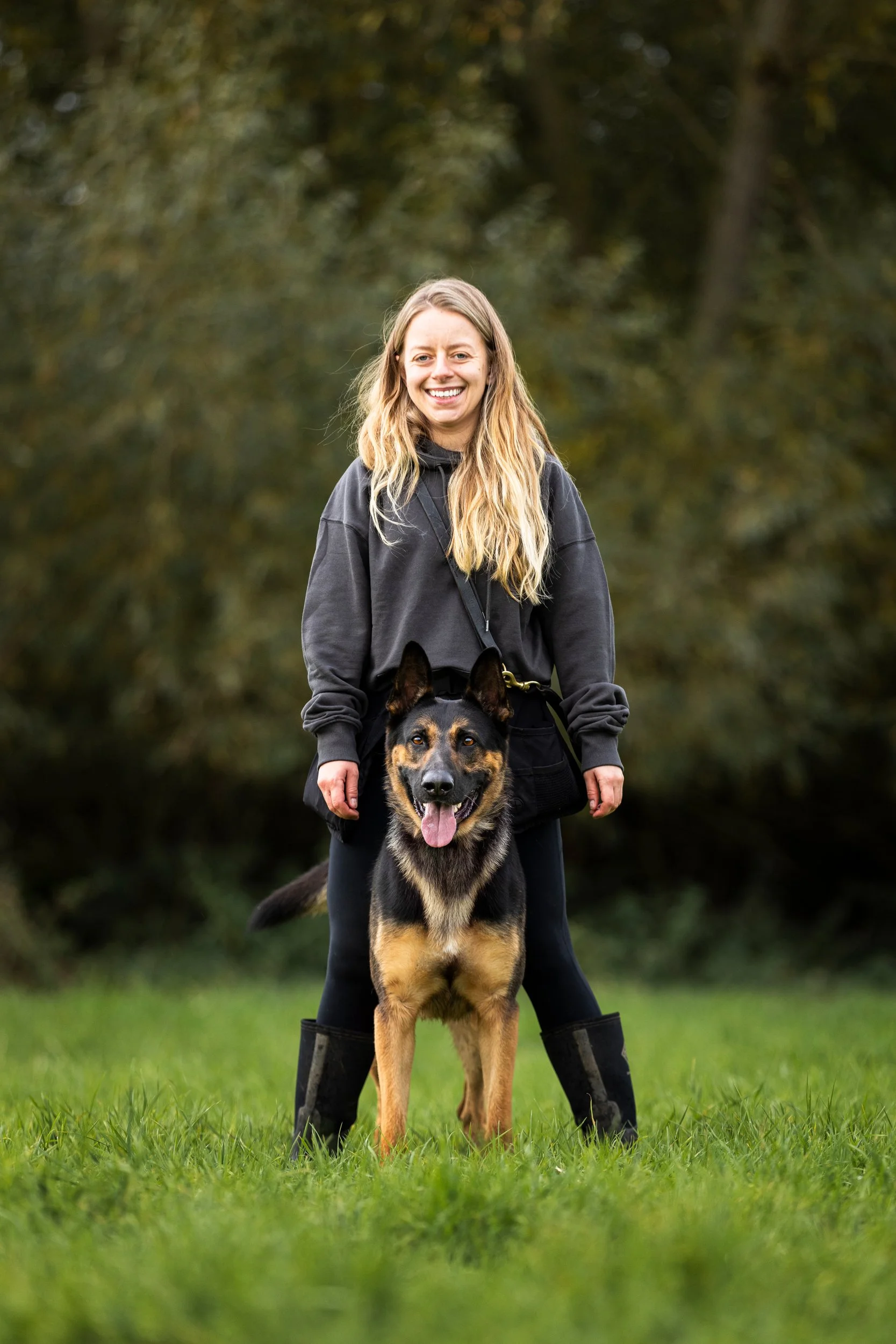 A woman in black pants and a gray hoodie standing outdoors on grass, smiling, with a black and tan shepherd dog sitting in front of her.