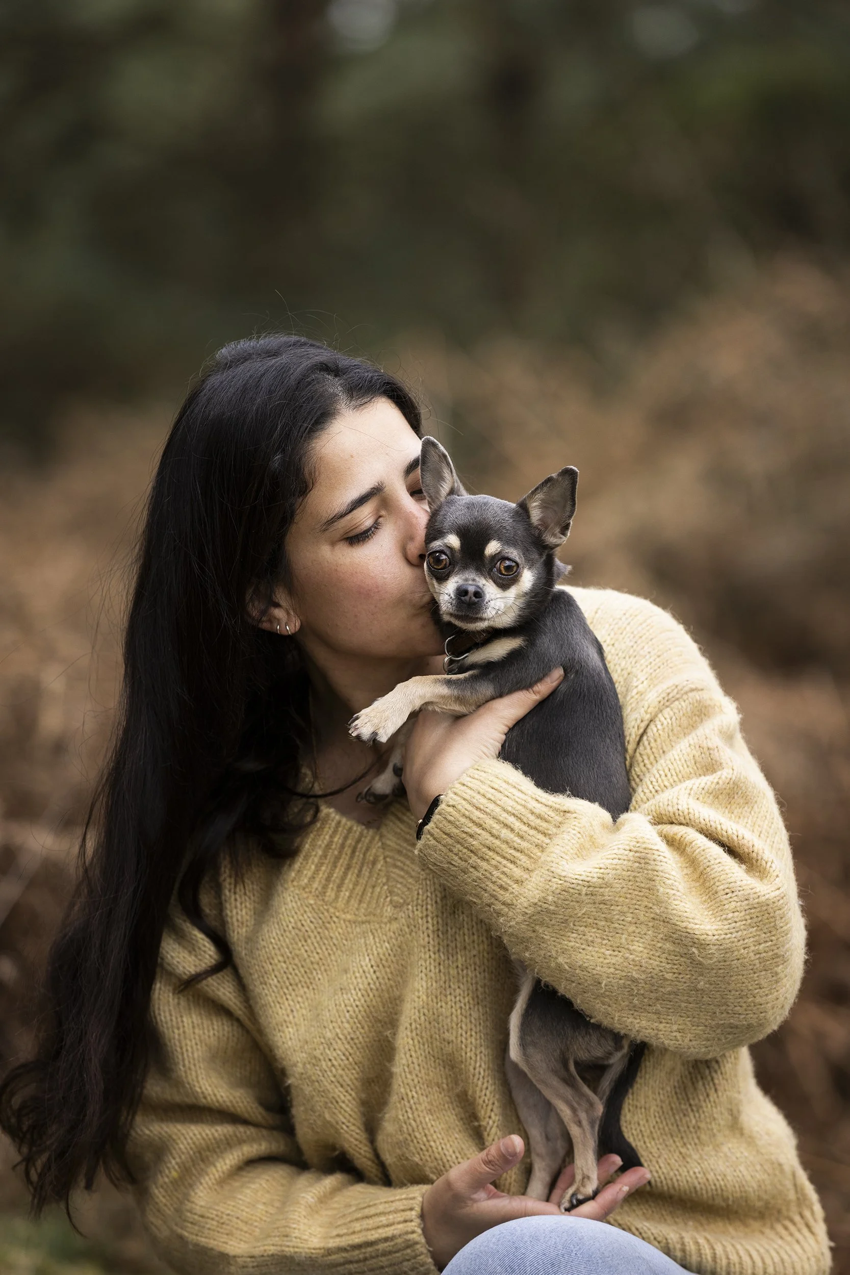 A woman with long dark hair wearing a beige sweater holding and kissing a small black and tan Chihuahua dog outdoors.