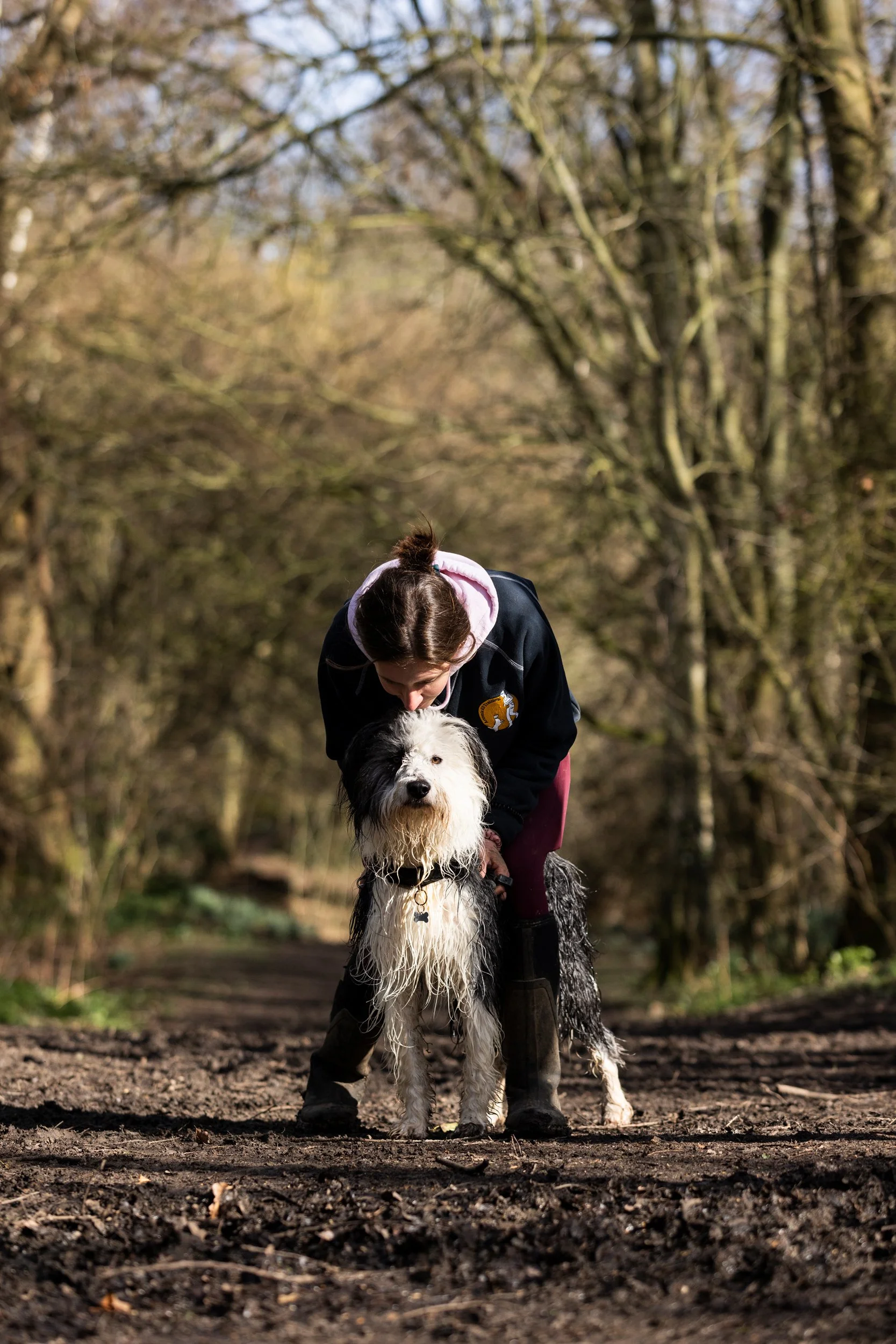 A young woman is bending down on a dirt trail in a forest, holding the leash of a black and white dog with a wet, shaggy coat.