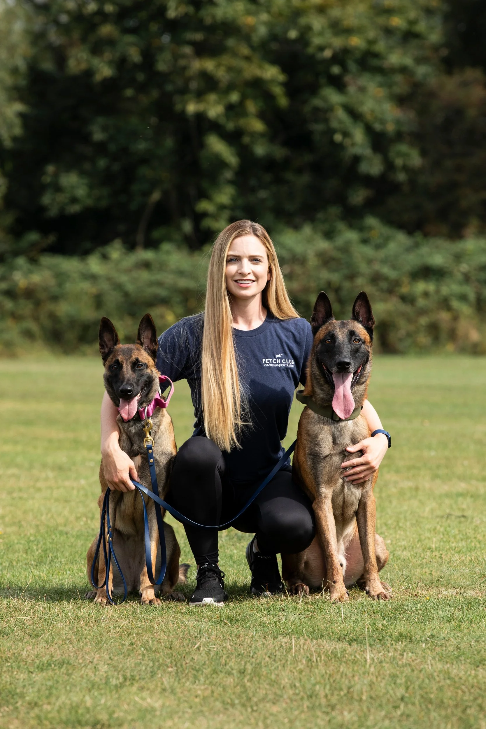 A woman kneeling on grass holding two Belgian Malinois dogs, one on each side, outdoors with trees in the background.