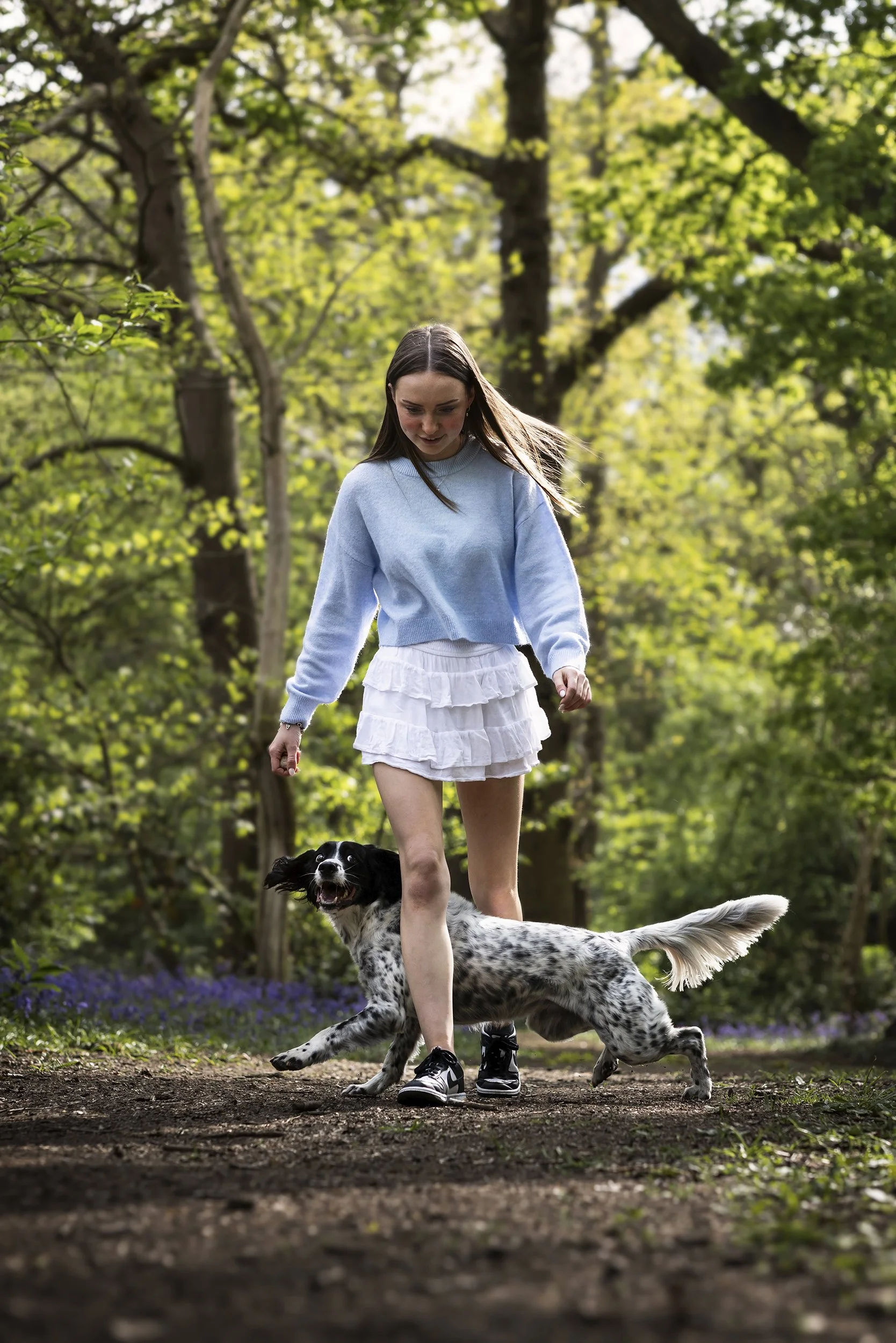 A young woman in a light blue sweater and white ruffled skirt walking with a black and white dog through a wooded park on a sunny day.