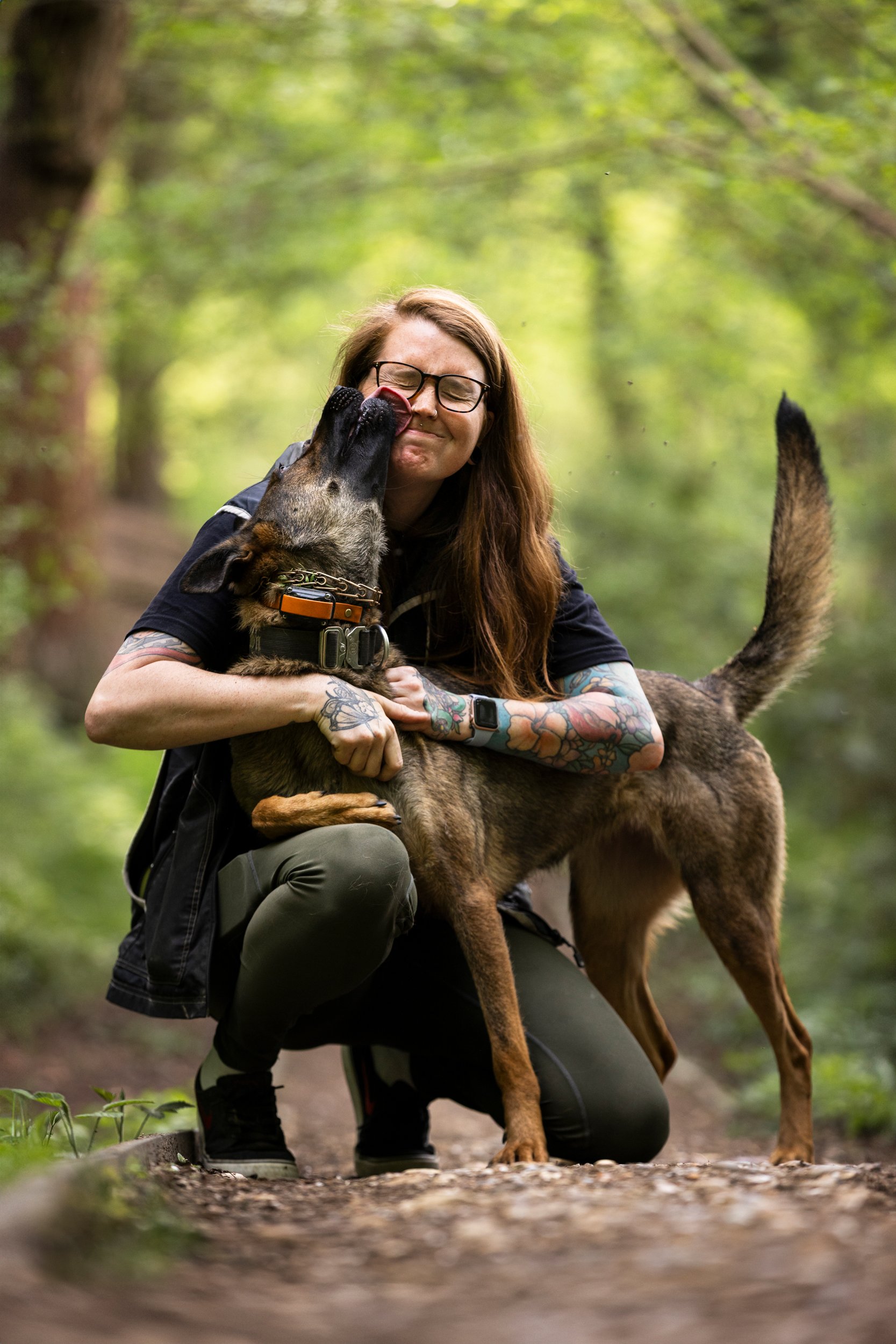 A woman with tattoos and glasses kneeling in a forest, hugging a Belgian Malinois dog as it licks her face.