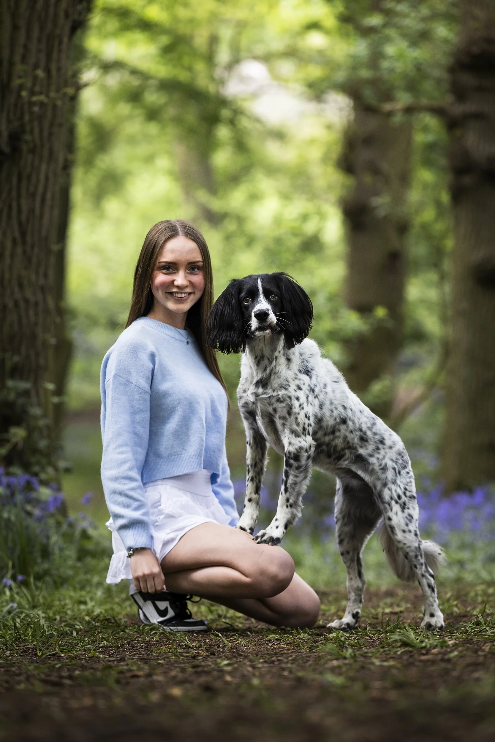A young woman in a blue sweater and white skirts kneeling on the ground beside a black and white dog in a forest during daytime.