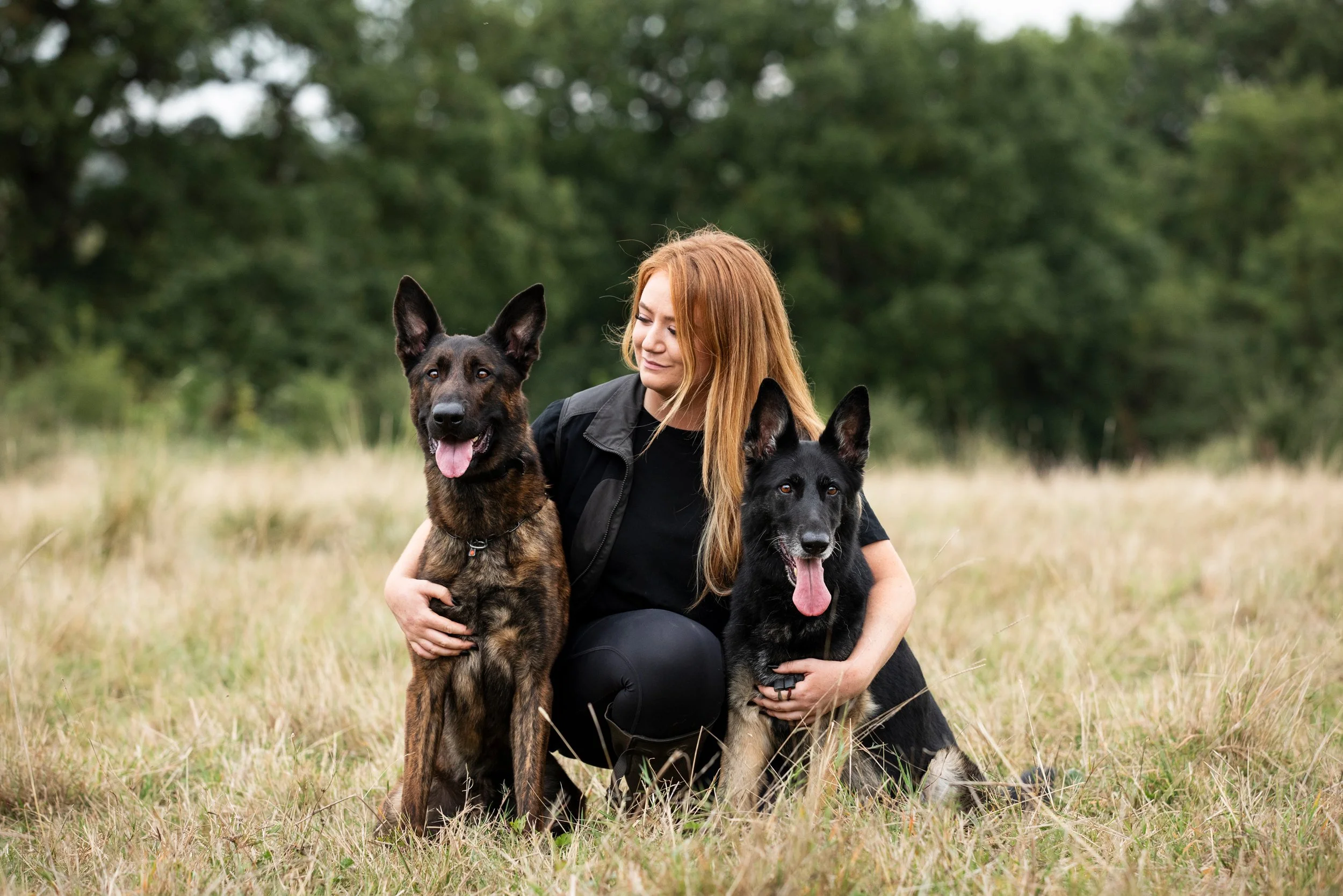 A woman with red hair kneels in a grassy field, holding two German Shepherd dogs, one on each side, with trees in the background.