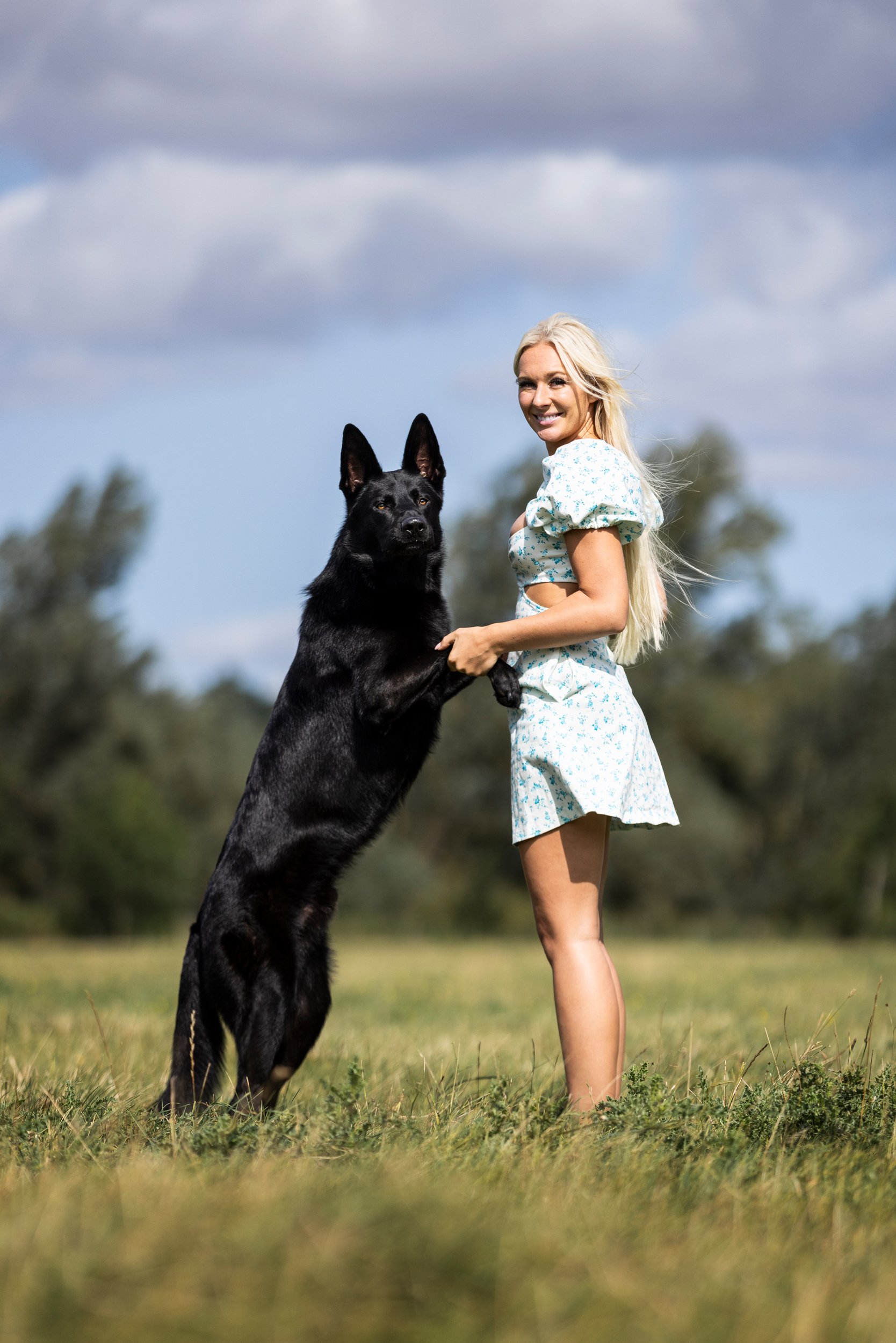 A woman in a blue floral dress holding a black Belgian Malinois dog standing on its hind legs in a grassy field with trees and a cloudy sky in the background.