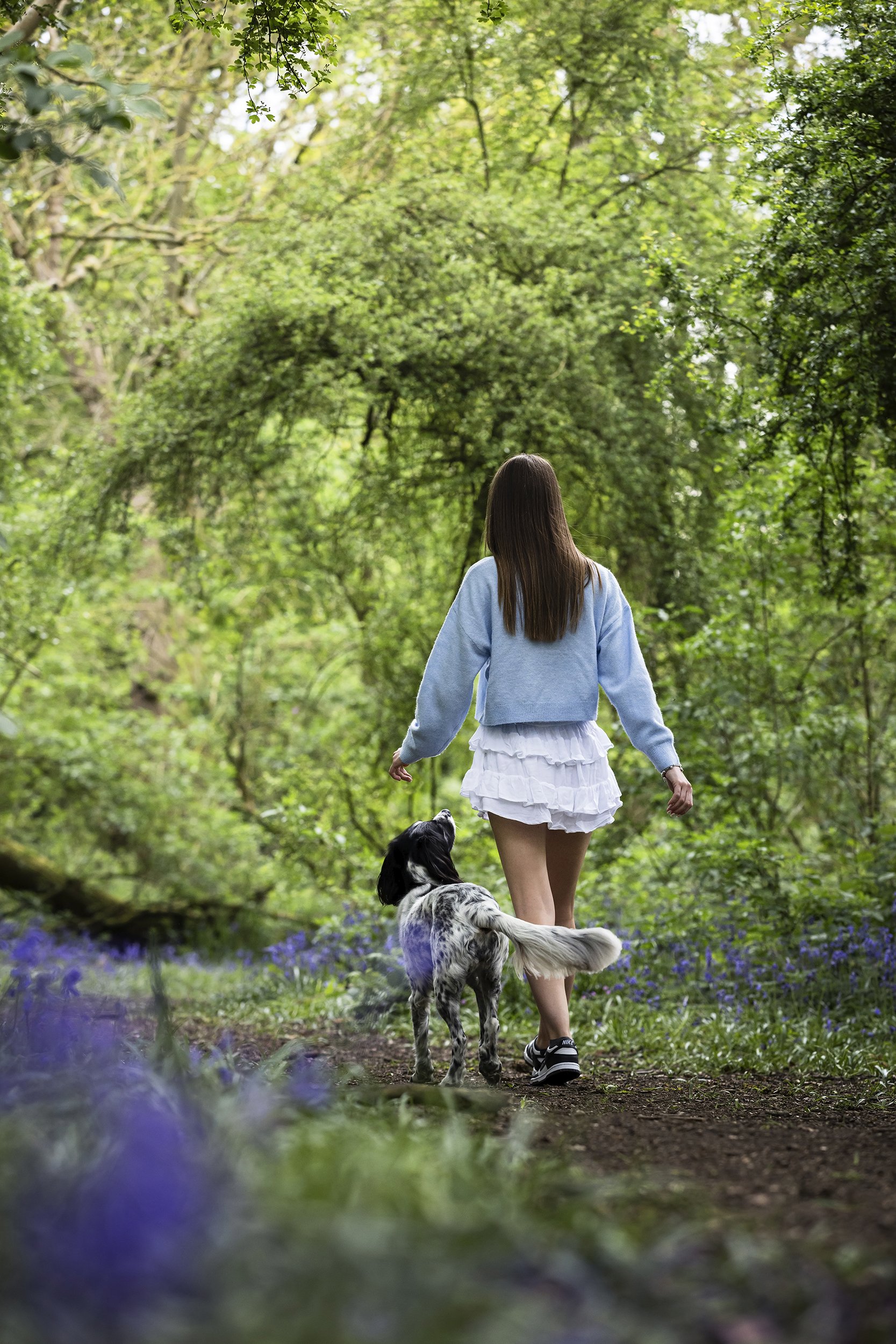 A woman in a light blue sweatshirt and white ruffled skirt walking with a black and white dog on a forest trail surrounded by green trees and purple flowers.