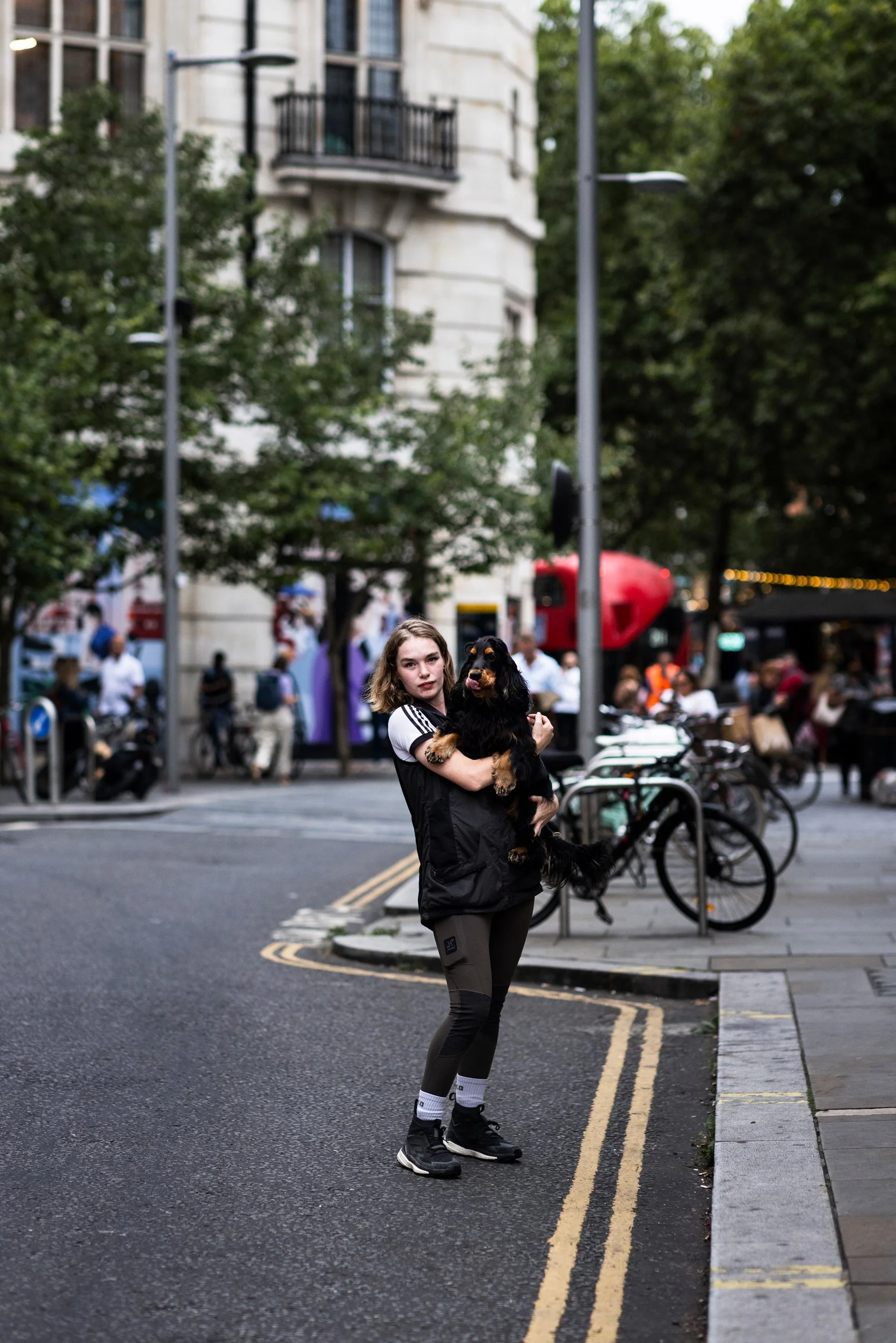 A young woman standing on a city street holding a large black and tan dog. There are bicycles parked nearby and a busy sidewalk with people and buildings in the background.