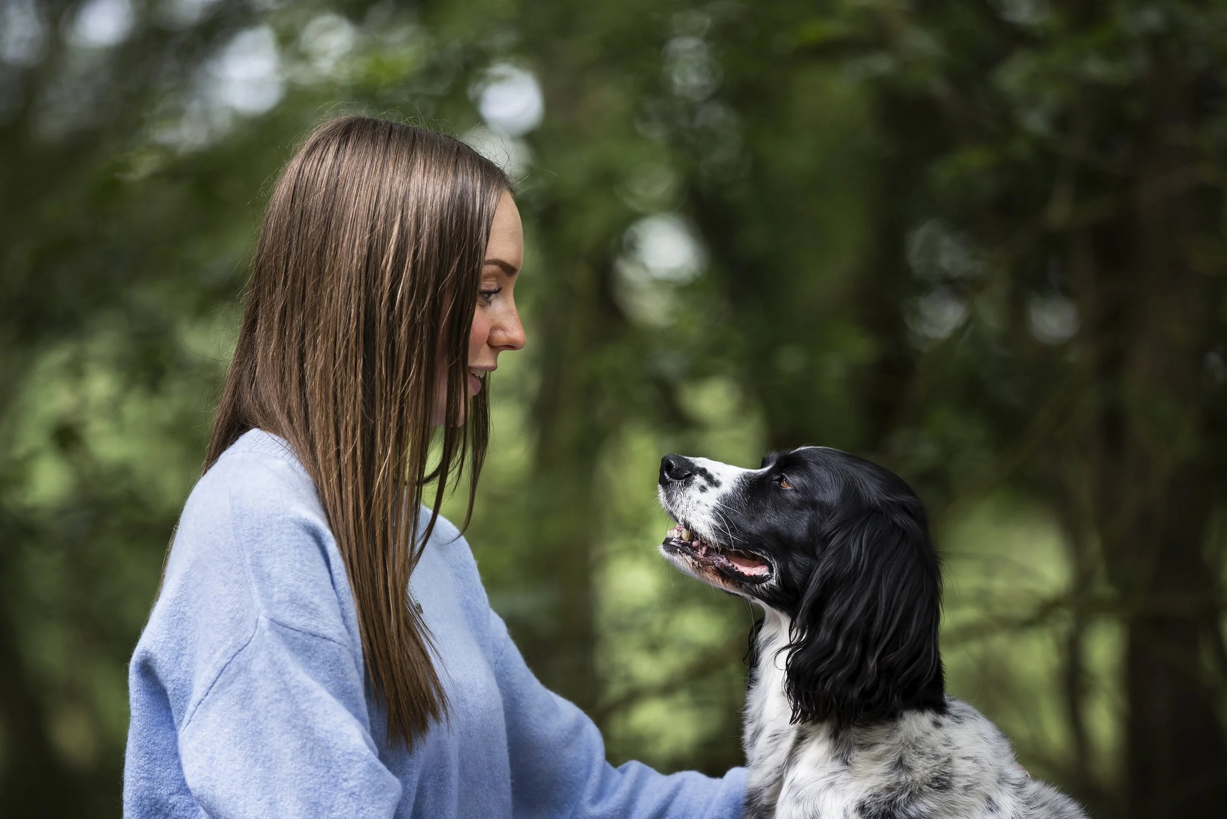 A woman and a black and white dog looking at each other in a wooded outdoor setting.