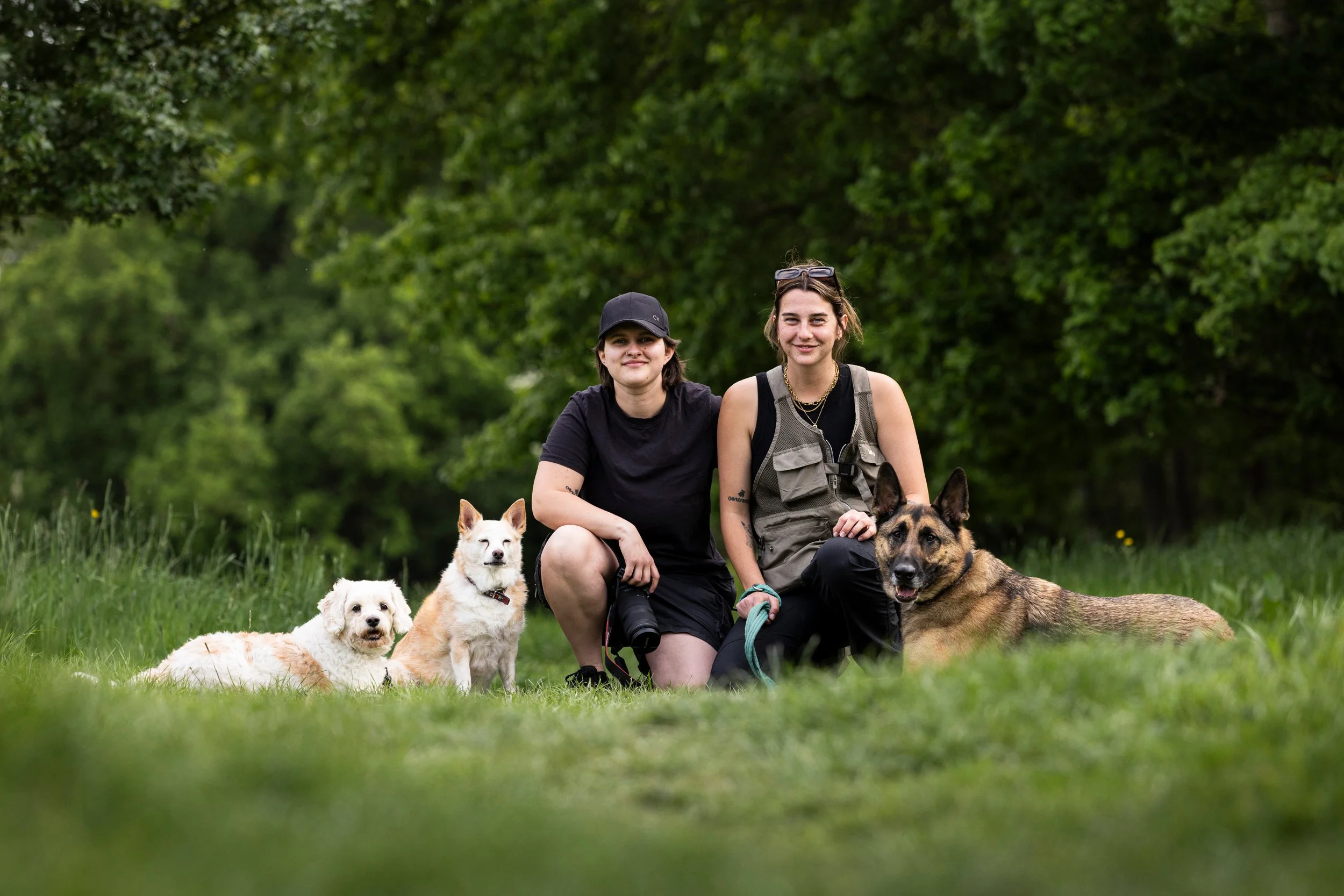 Two women and four dogs sitting on grass in a park with green trees in the background.