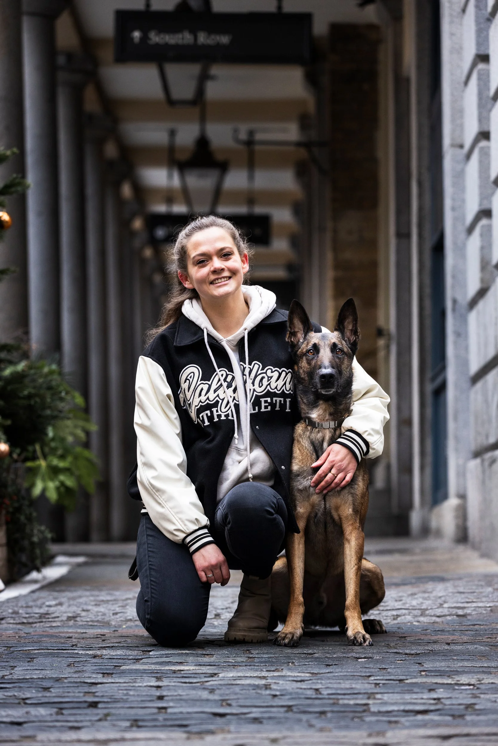 A young woman smiling and kneeling next to a German Shepherd dog in an outdoor alleyway, with a sign indicating 'South Row' visible overhead.