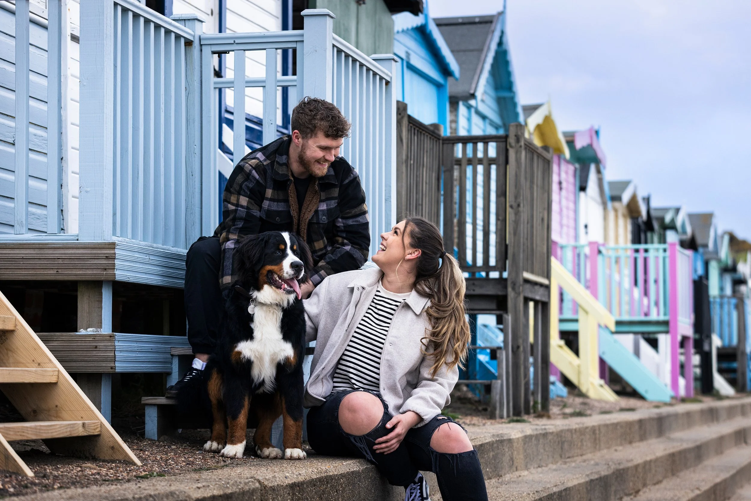A couple and their dog near colorful beach houses. The man is on the stairs, leaning towards the woman, who is sitting on the sidewalk with her hand on the dog. They are smiling at each other.