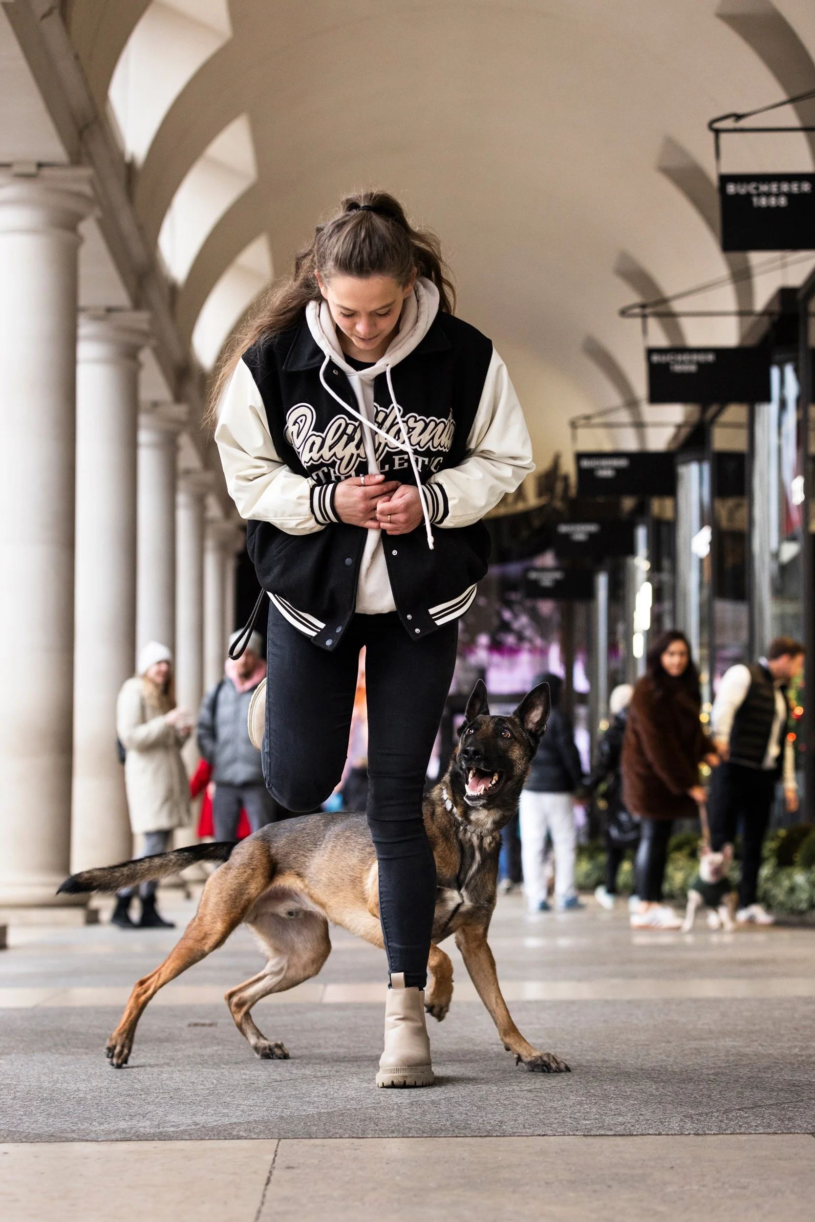 A young woman with long hair in a ponytail, wearing a black and white varsity jacket, black pants, and beige boots, is walking with a Belgian Malinois dog on a leash in a covered shopping plaza. The woman is looking down at the dog, which appears hap
