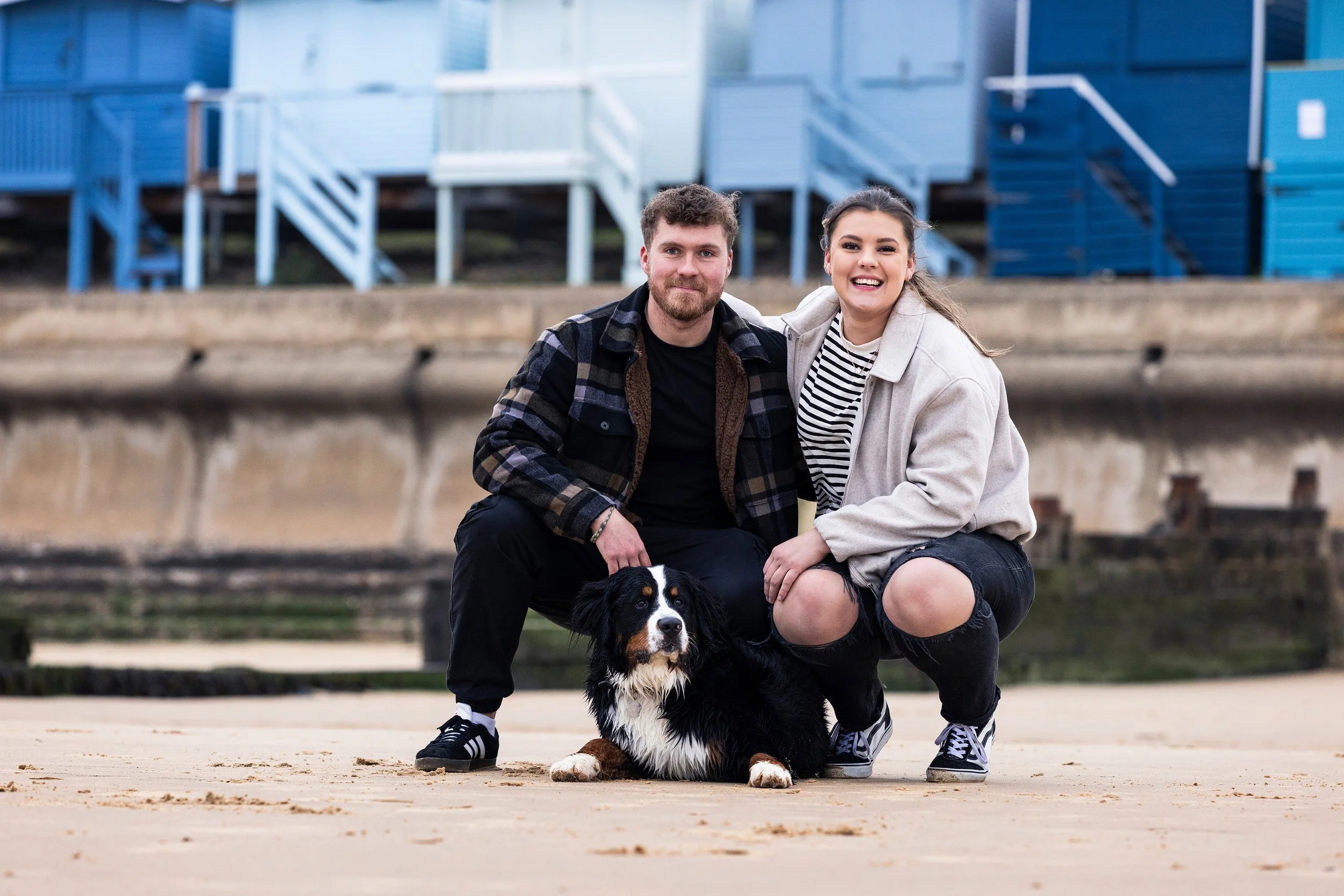 A young man and woman kneeling on a sandy beach with a black, white, and brown dog in front of them, with blue houses in the background.