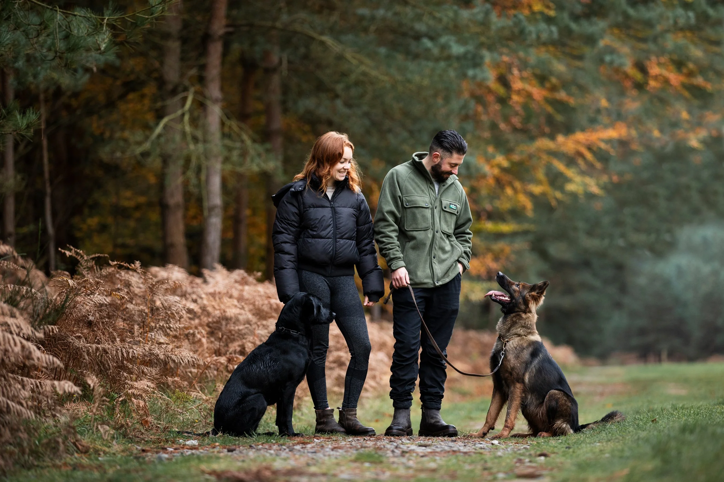 A man and a woman walking with two dogs on a forest trail during autumn. The woman has red hair and wears a black jacket, while the man has dark hair and a beard, wearing an olive green jacket. The black dog sits beside the woman, and a German Shephe