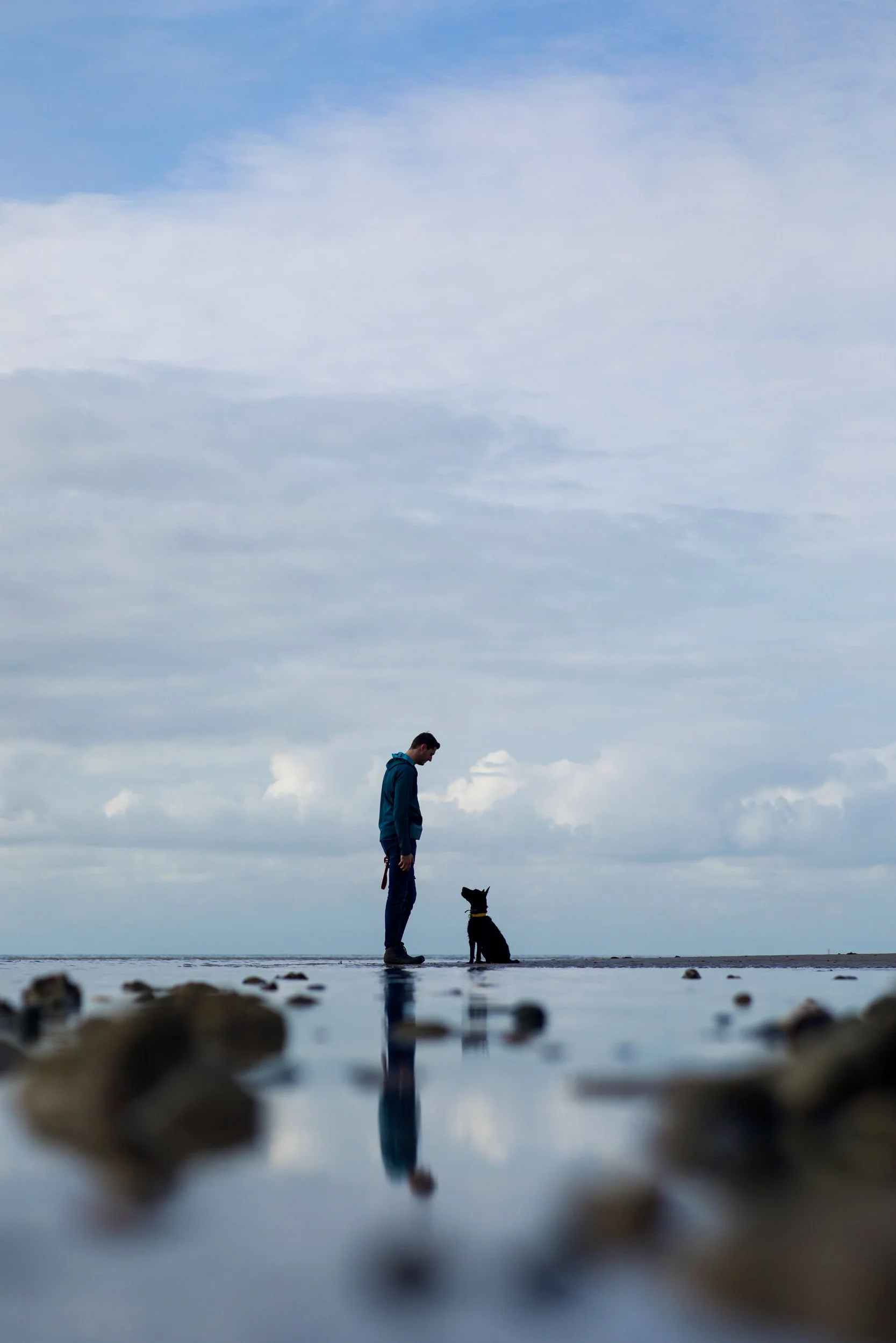 A person and a dog standing on a beach, reflected in a shallow pool of water, with a cloudy sky in the background.