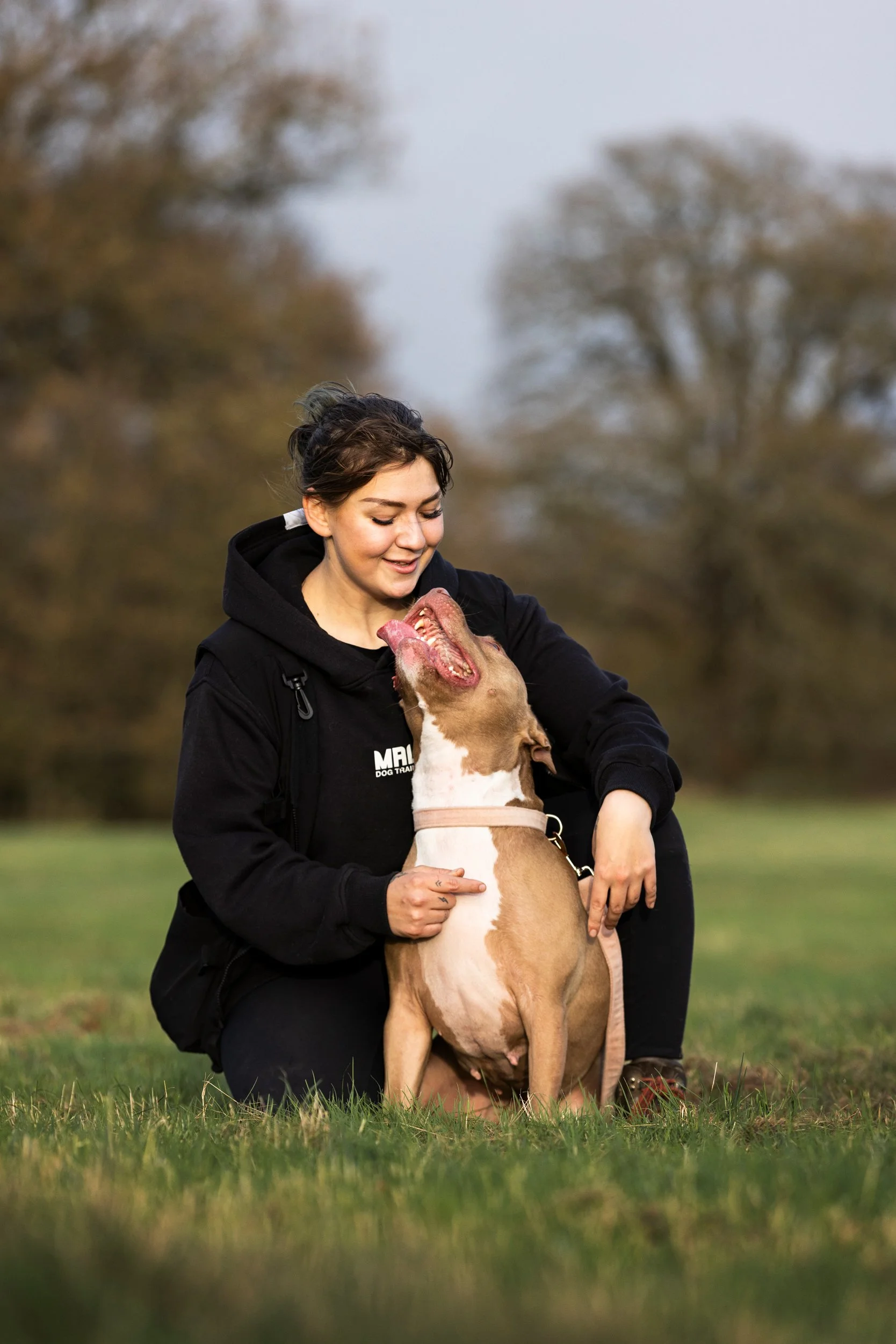 A young woman in a black hoodie kneeling on grass, smiling, with a brown and white dog sitting in front of her. The dog has its head tilted back and mouth open, looking happy.