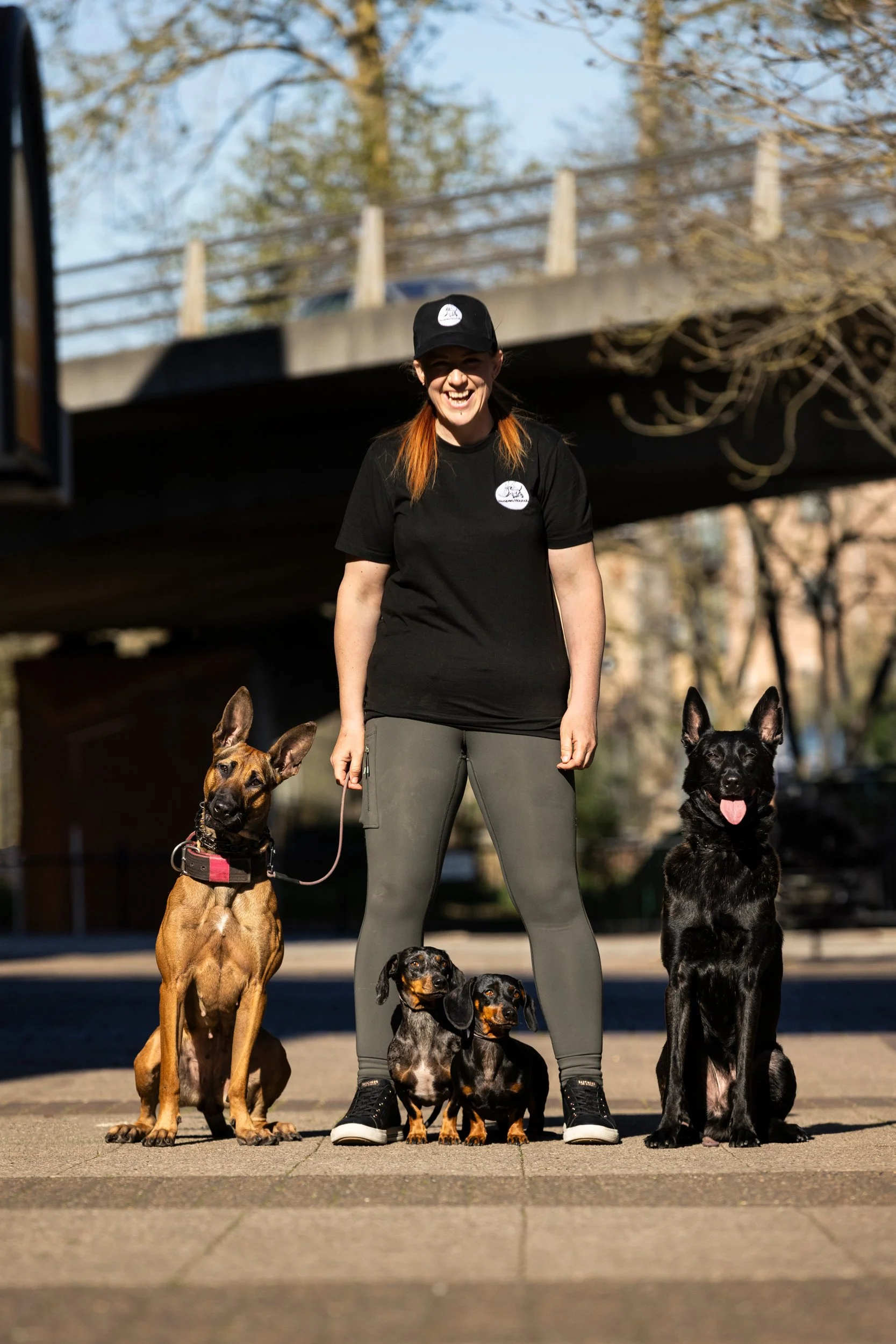 A woman standing outdoors under a bridge, smiling, with five dogs of various breeds sitting beside her on a paved surface with trees in the background.