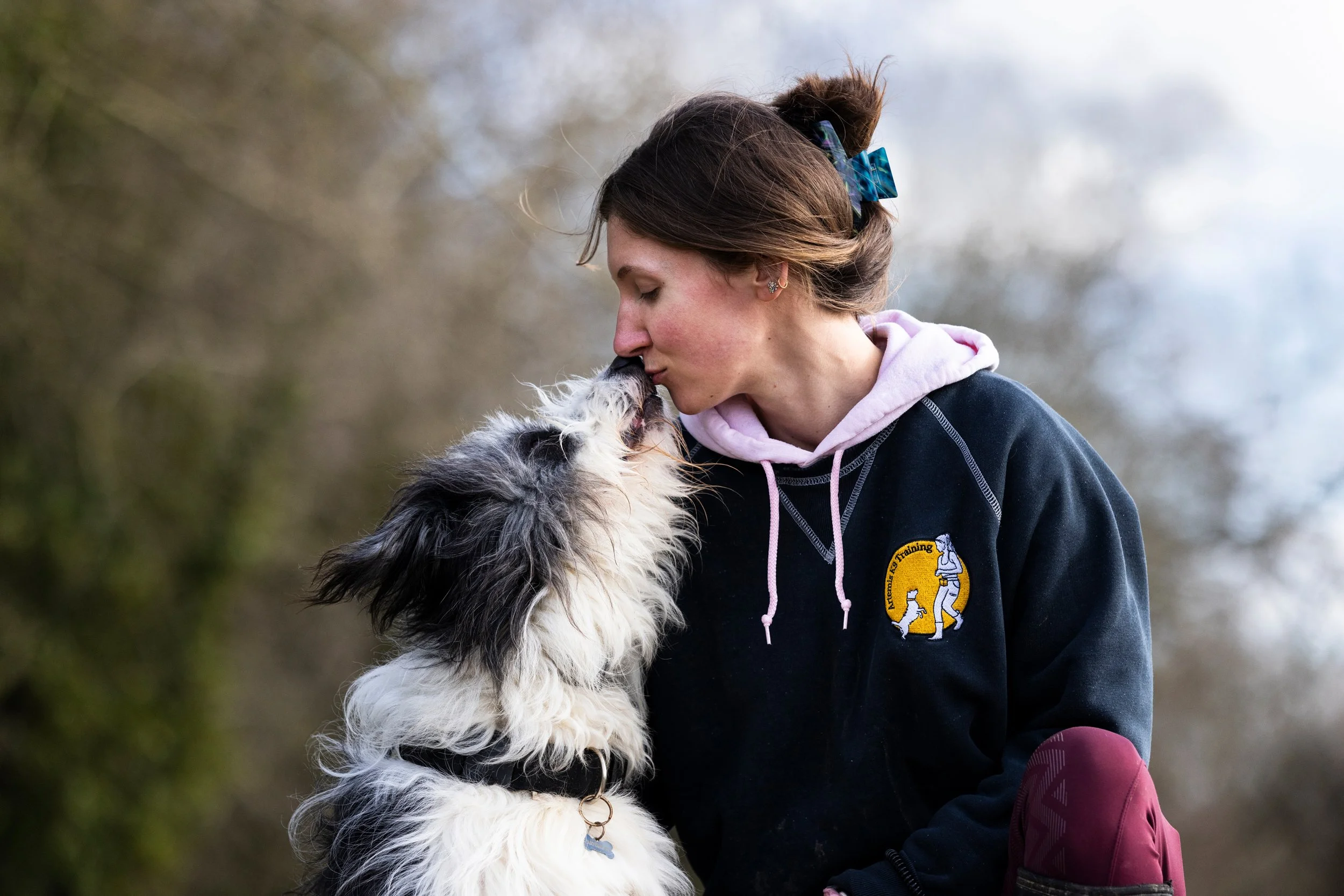 A young woman with brown hair tied back with a blue bow, wearing a black hoodie with a patch of a dog and the words 'K-9 Training,' is nose to nose with a black and white dog outdoors in a natural setting with blurred trees in the background.