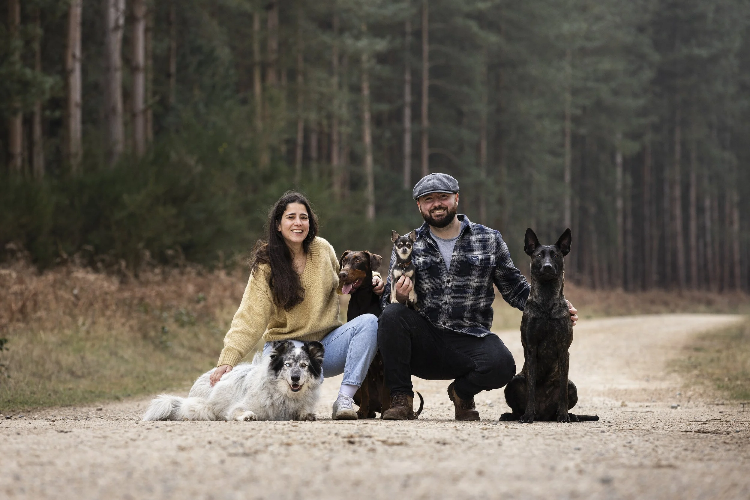 A smiling woman and man with four dogs on a forest dirt path.