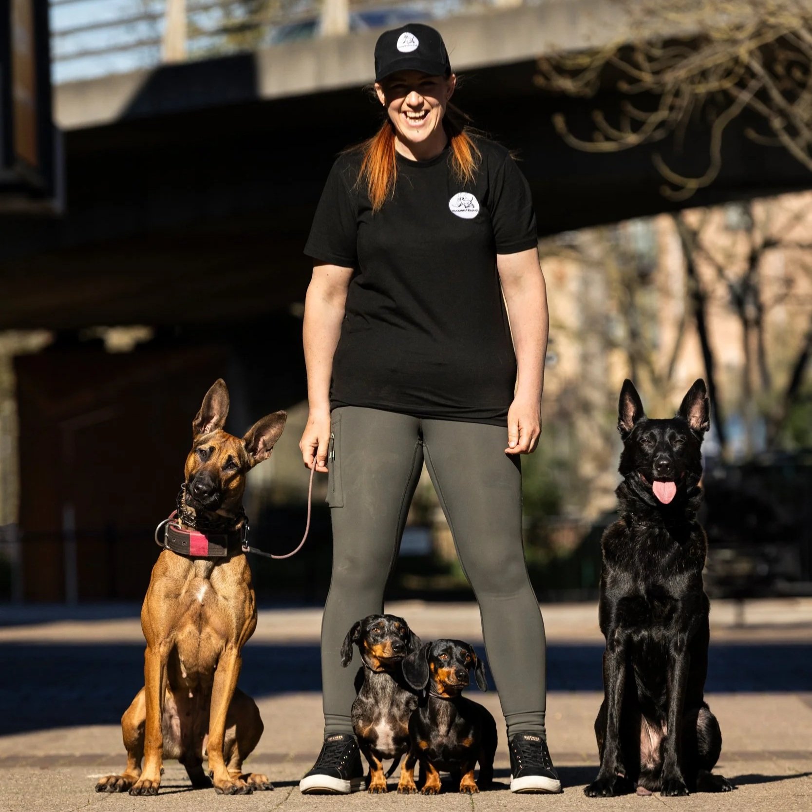A woman standing outdoors with five dogs of various breeds and sizes, smiling and holding their leashes.
