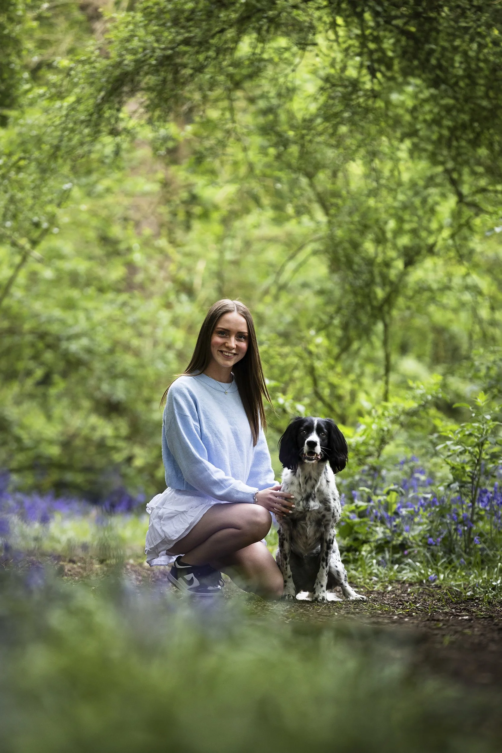 A young woman with long brown hair wearing a light blue sweater and white skirt kneels beside a black and white dog on a forest trail surrounded by green trees and purple wildflowers.