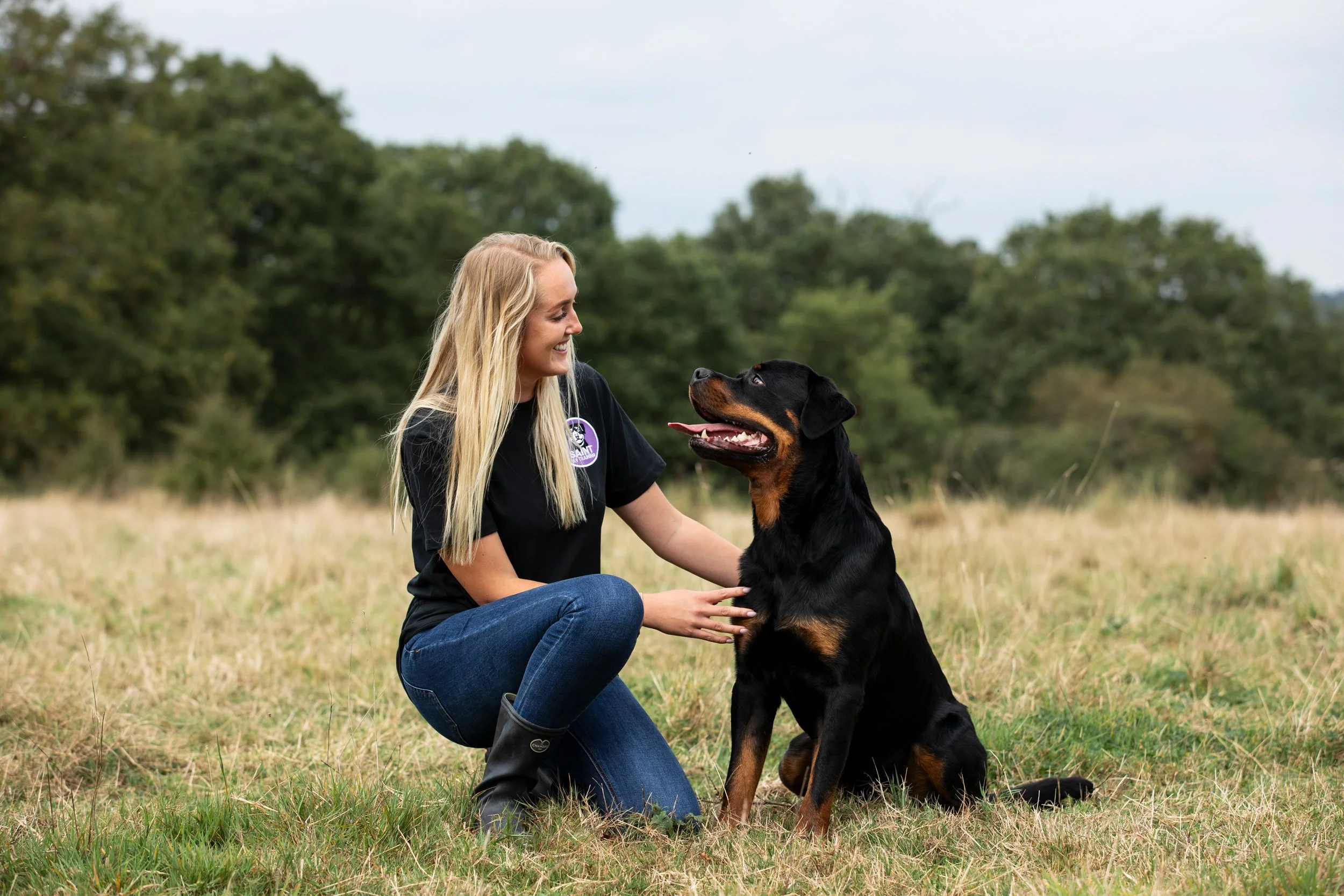A woman with long blonde hair kneeling on grass, smiling at a large black and tan Rottweiler sitting in front of her outdoors in a grassy field with trees in the background.