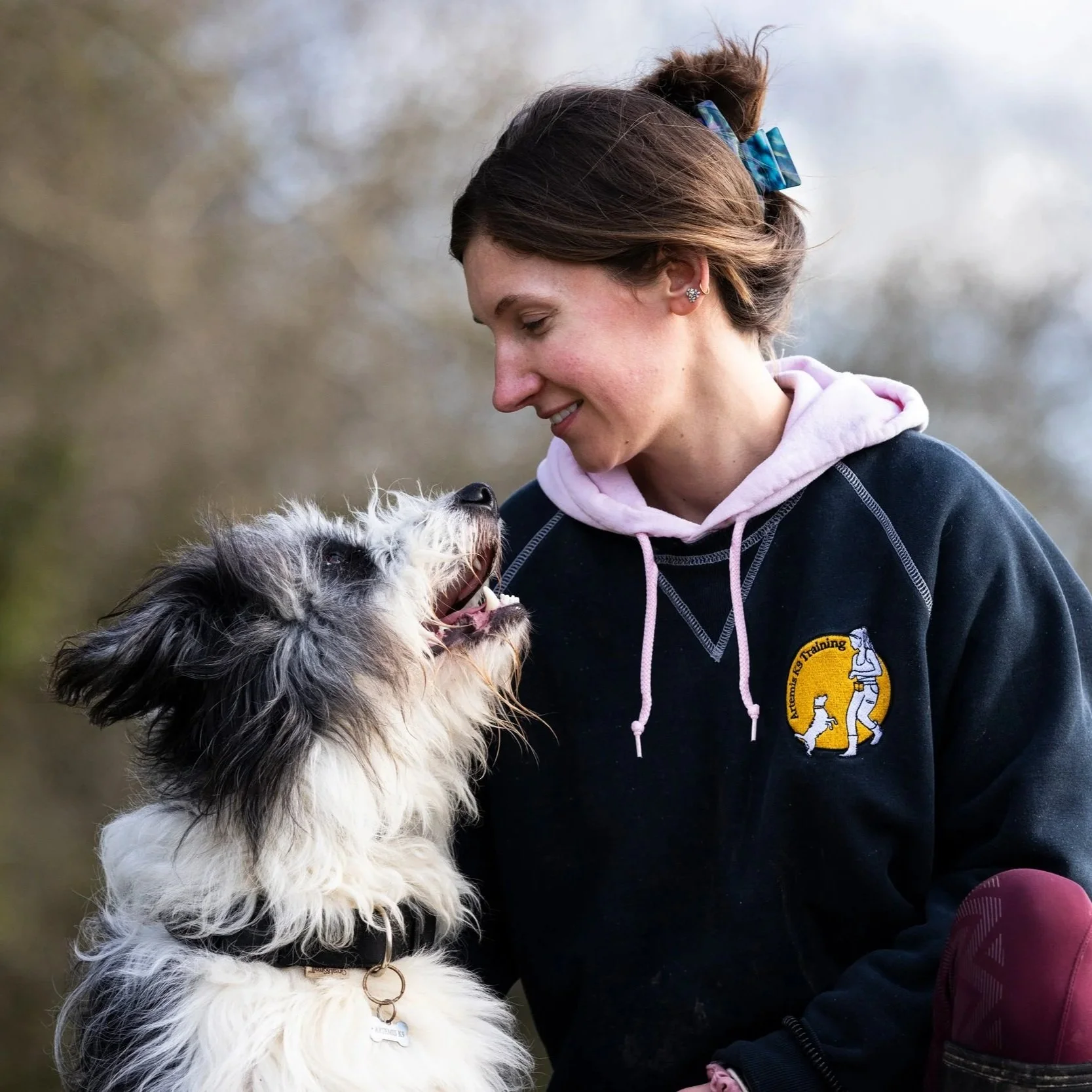 A woman with brown hair tied with a blue checkered ribbon, smiling, looking at a black and white dog affectionately. She is wearing a black hoodie with a yellow emblem, and the background is outdoors with blurred trees.
