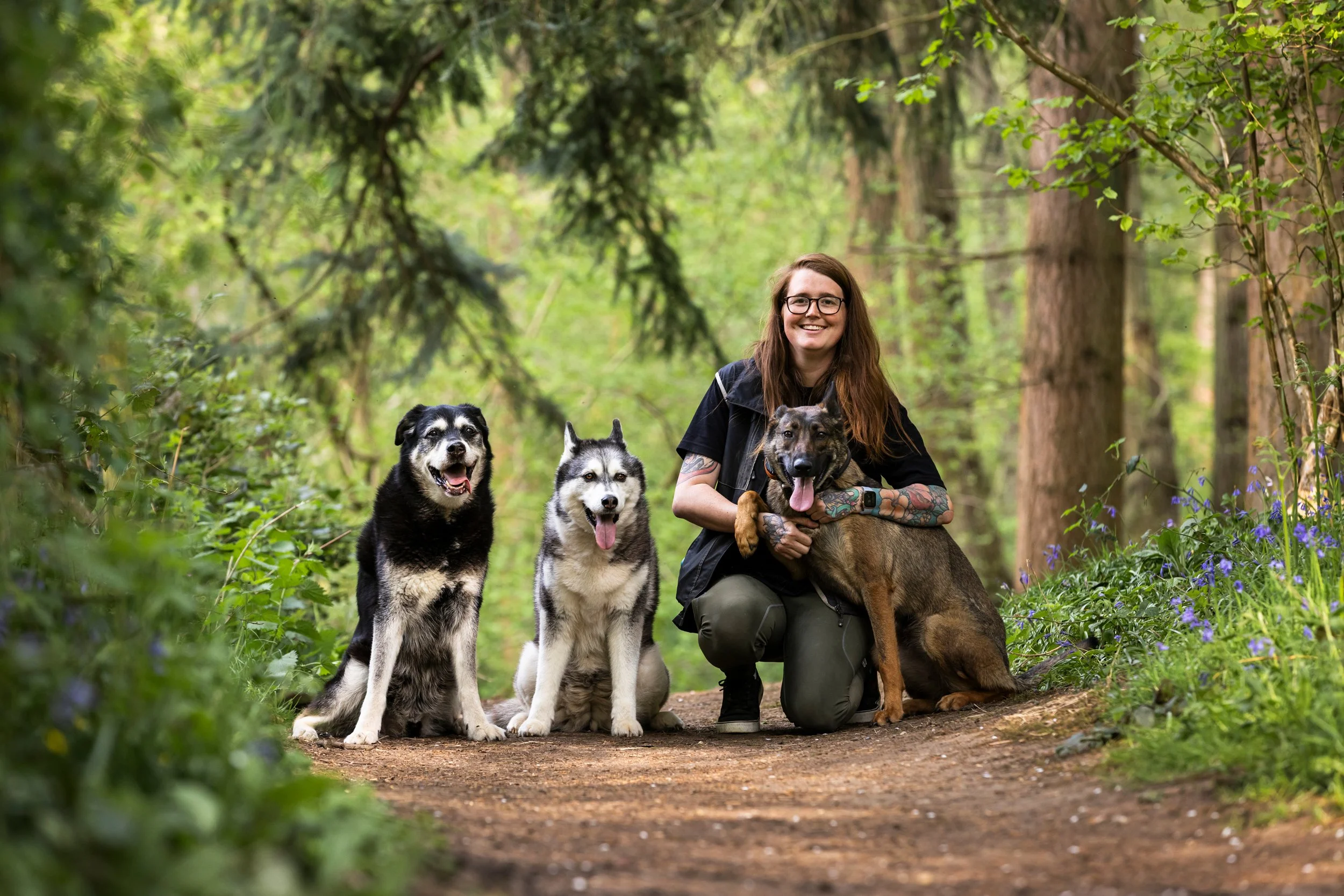 A woman with tattoos, glasses, and long brown hair kneeling on a forest trail next to three dogs, two sitting and one standing, with tall trees and purple flowers surrounding them.