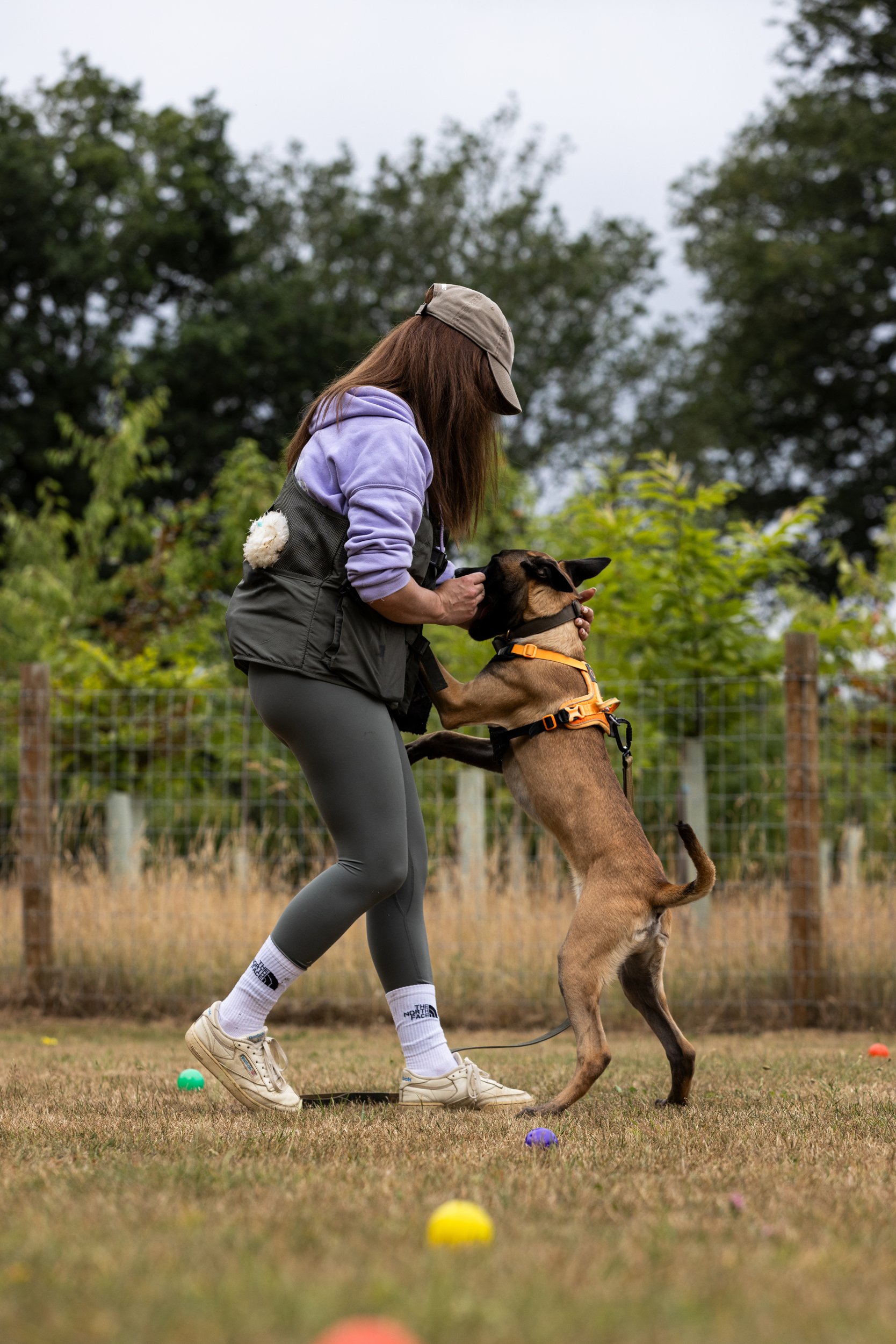 A woman in athletic clothing is playing with a dog outdoors in a grassy area with colorful balls around them.