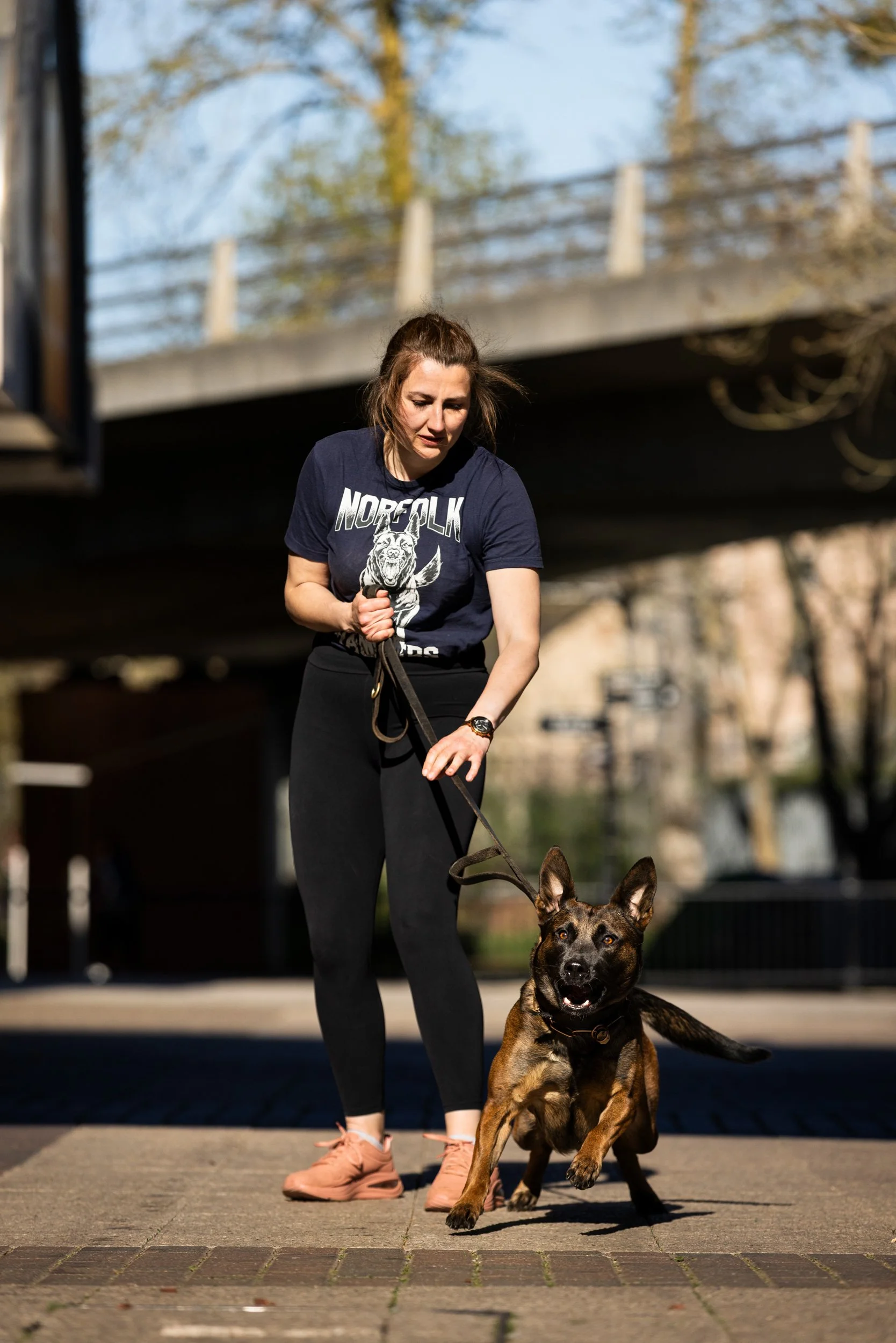 A woman in black leggings and a navy graphic T-shirt is walking a brown and black dog on a leash in an outdoor city area under a bridge.