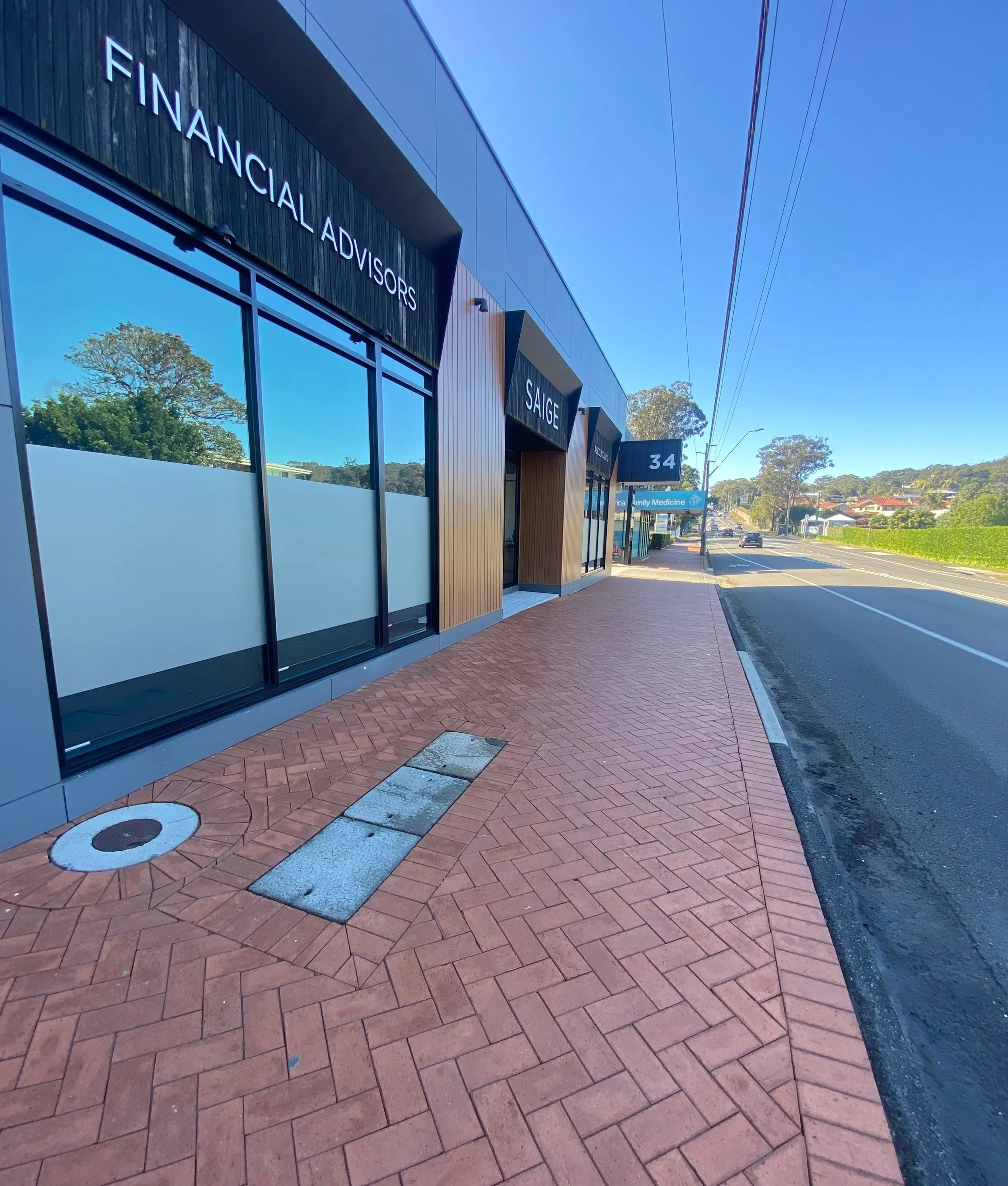 Exterior view of a modern commercial building with signs 'Financial Advisors' and 'Saige' on the facade, along a sidewalk with a brick pattern. The street is visible on the right with some cars and houses in the background, and the sky is clear with power lines overhead.