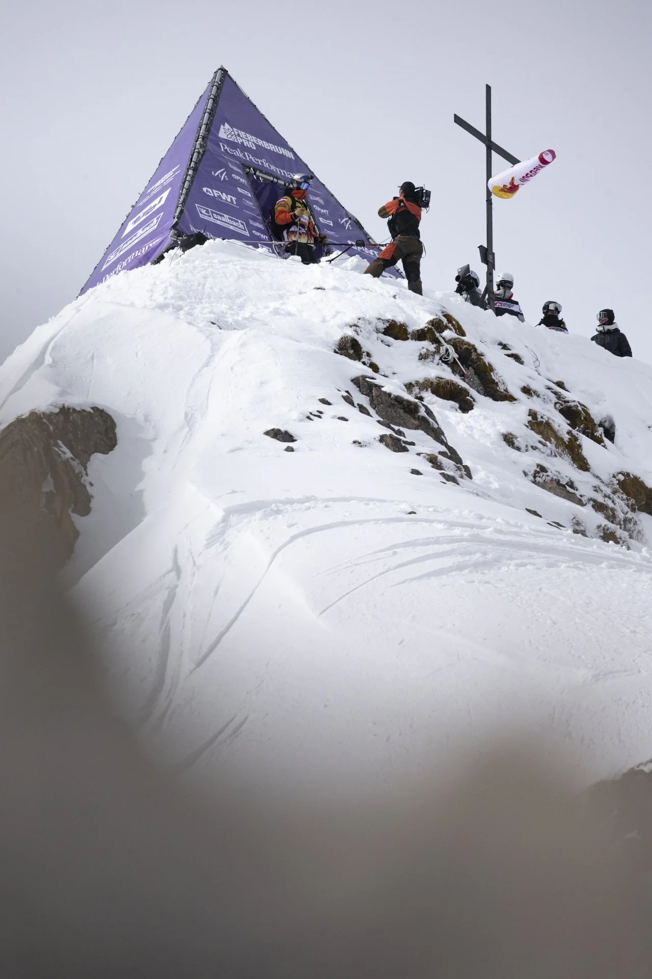 Verschneiter Berggipfel mit einem großen Kreuz und mehreren Menschen, die auf den Berg vorbereitet zu sein scheinen. Ein violettes Banner mit Logos ist ebenfalls sichtbar. Ein Kameramann nimmt das Geschehen auf.