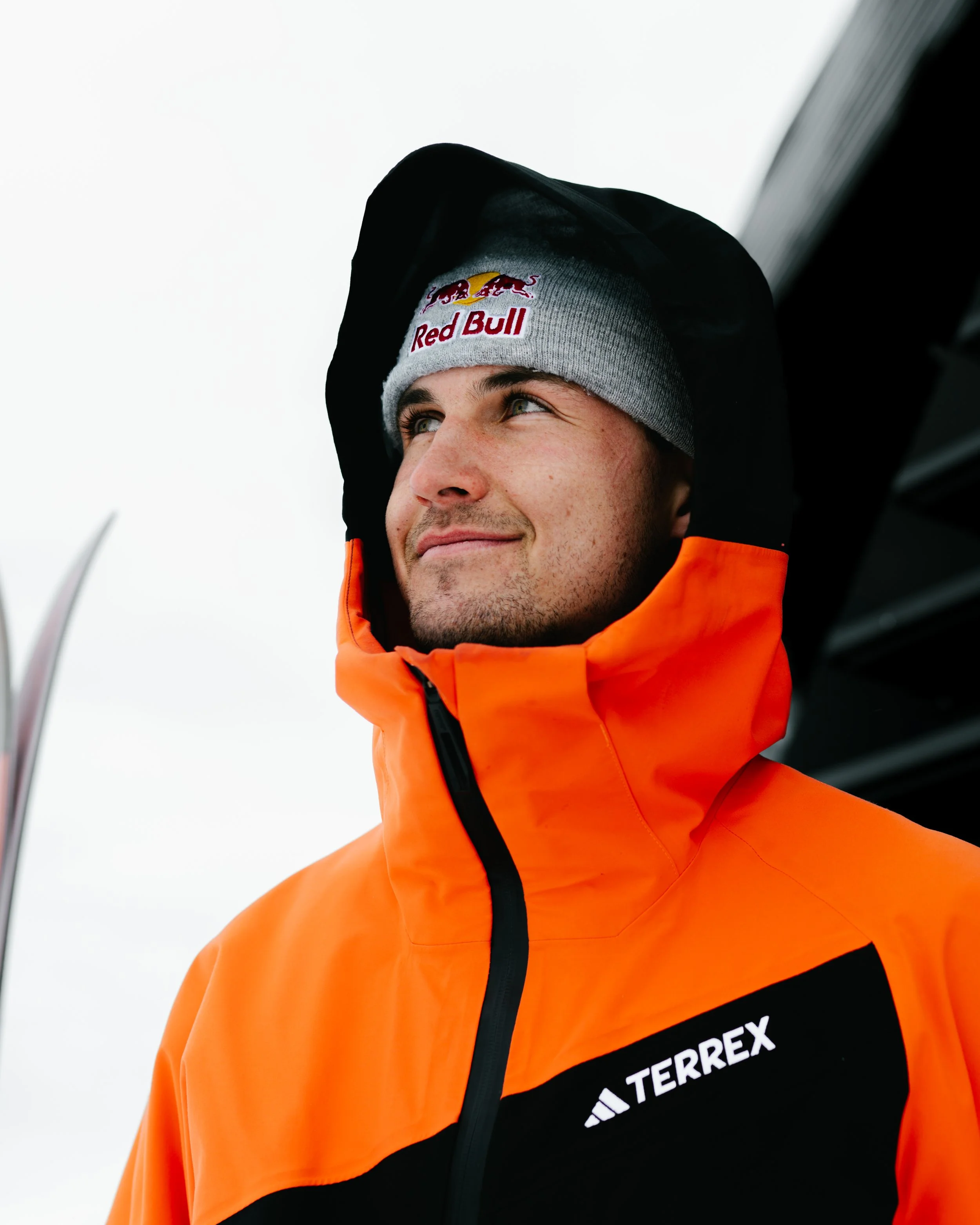 Mann in oranger Jacke mit 'TERREX'-Logo, trägt eine graue Mütze mit 'Red Bull'-Logo, schaut nach oben.