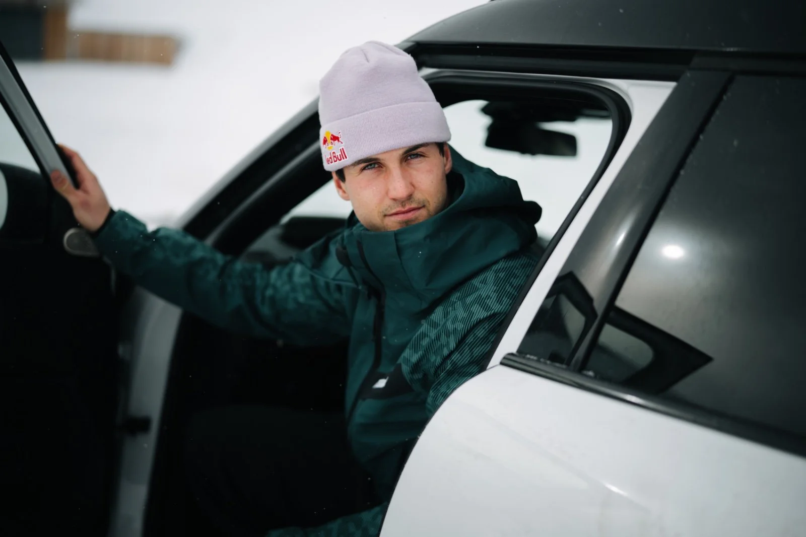 Mann mit Mütze in winterlicher Kleidung, sitzt in einem Auto mit geöffneter Tür in schneebedeckter Umgebung.